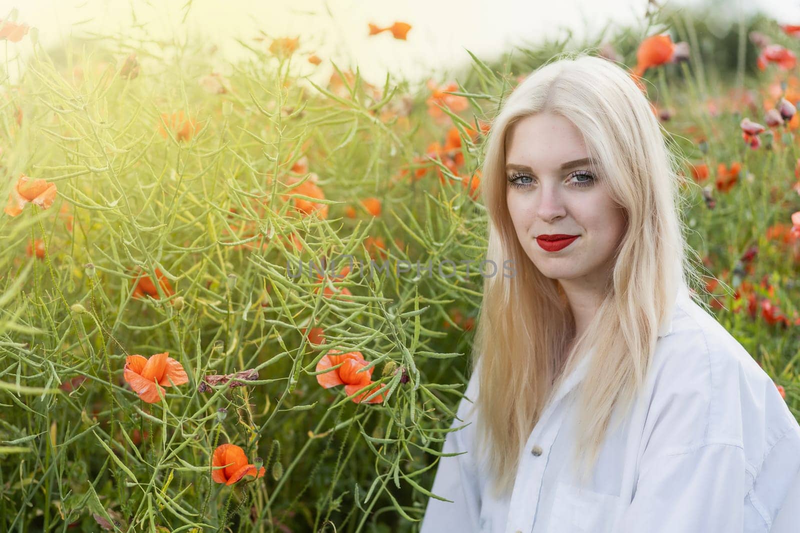 Portrait of blonde young woman posing in red poppy field smiling at the camera. Horizontally.
