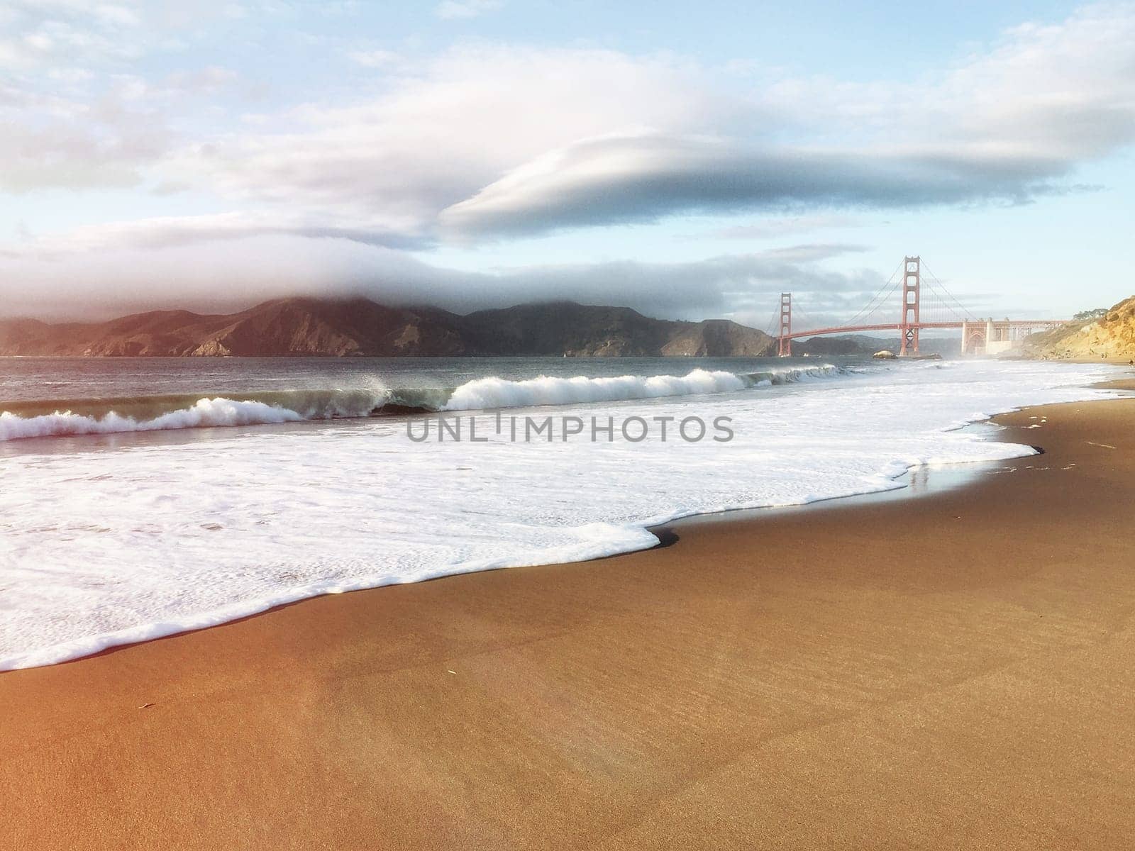 Golden gate bridge view from beach. Cloudy sky in background, waves in foreground.