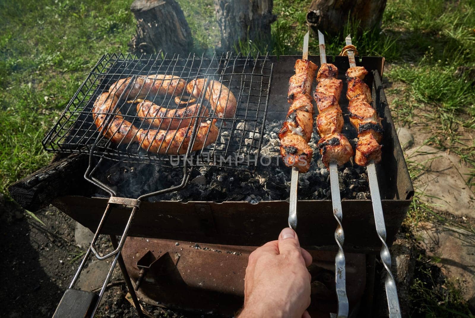 Man preparing shashlik or shish kebab over charcoal. Barbeque meat and sausages or bratwurst on a grill grate in backyard. Grilled meat on metal skewer outdoor. BBQ party or picnic food. Close up shot