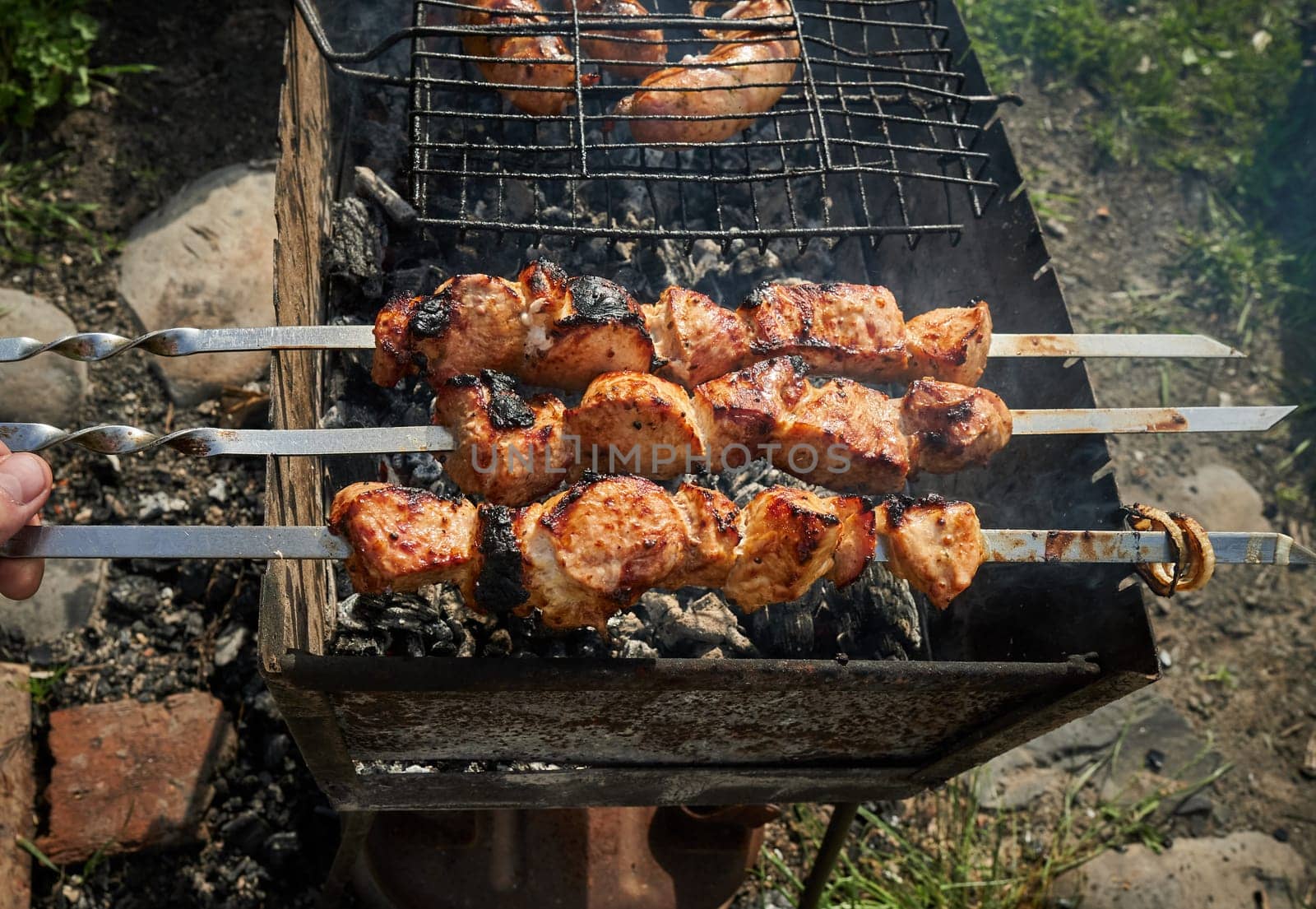 Man preparing shashlik or shish kebab over charcoal. Barbeque meat and sausages or bratwurst on a grill grate in backyard. Grilled meat on metal skewer outdoor. BBQ party or picnic food. Close up shot