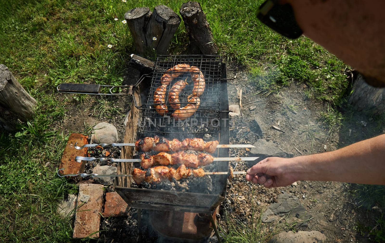 Man preparing shashlik or shish kebab over charcoal. Barbeque meat and sausages or bratwurst on a grill grate in backyard. Grilled meat on metal skewer outdoor. BBQ party or picnic food. Close up shot