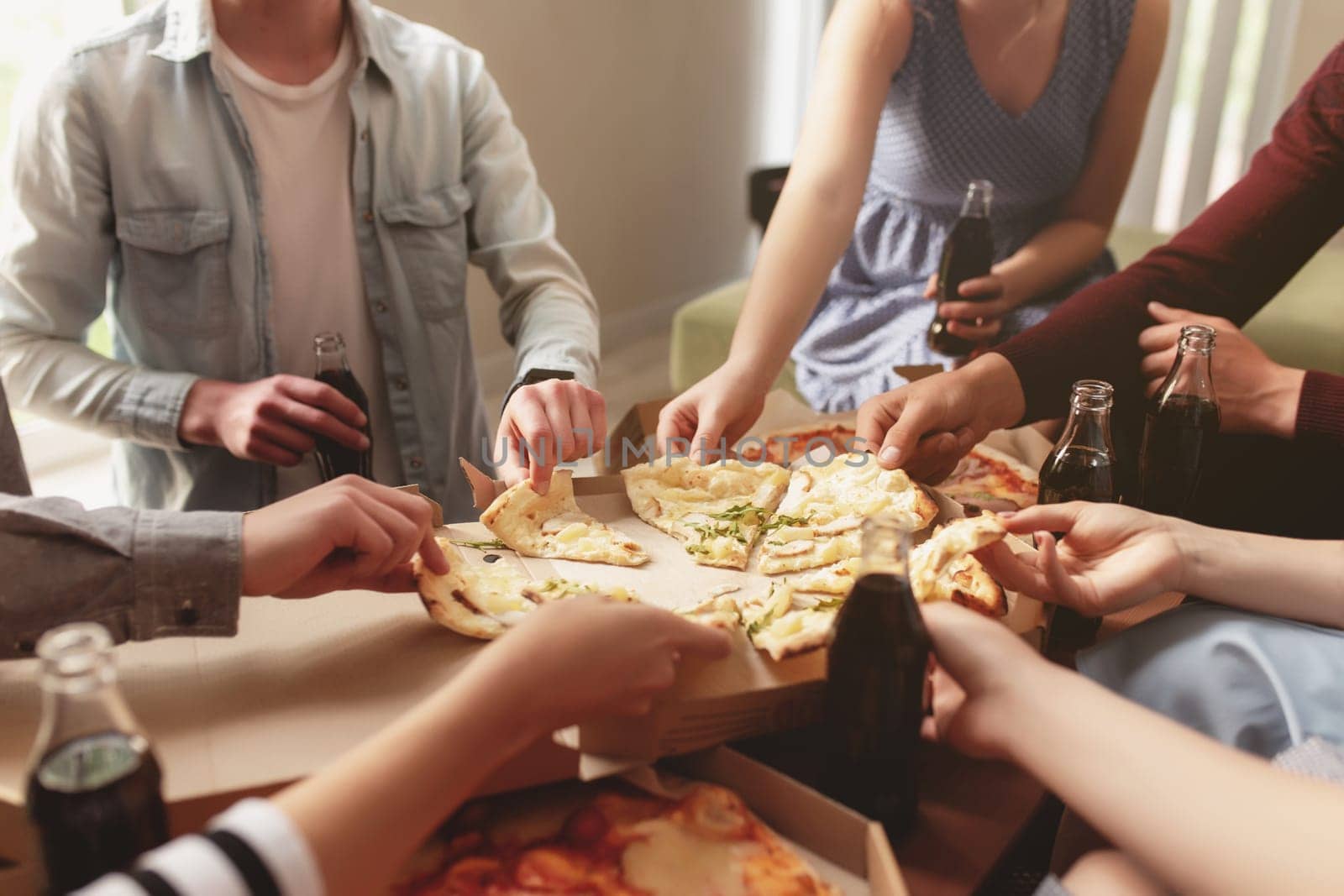 A pizza party, a group of friends chatting and happily eating pizza at the table and drinking sweet soda water. Many friends handing out together because of birthday celebration. Cheeses pizza pieces