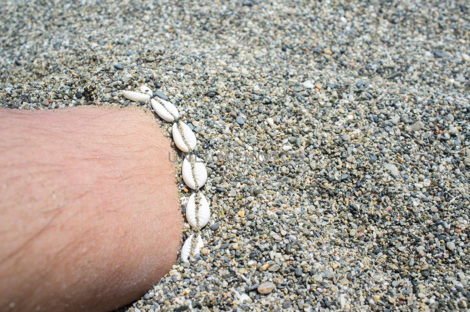 Shell ankle bracelet on half buried male leg. Symbol of summertime