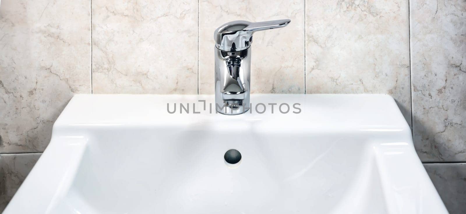 Chromed faucet with lever to mix hot and cold water on a modern white sink in a bathroom with tiled walls. Domestic life and hygiene