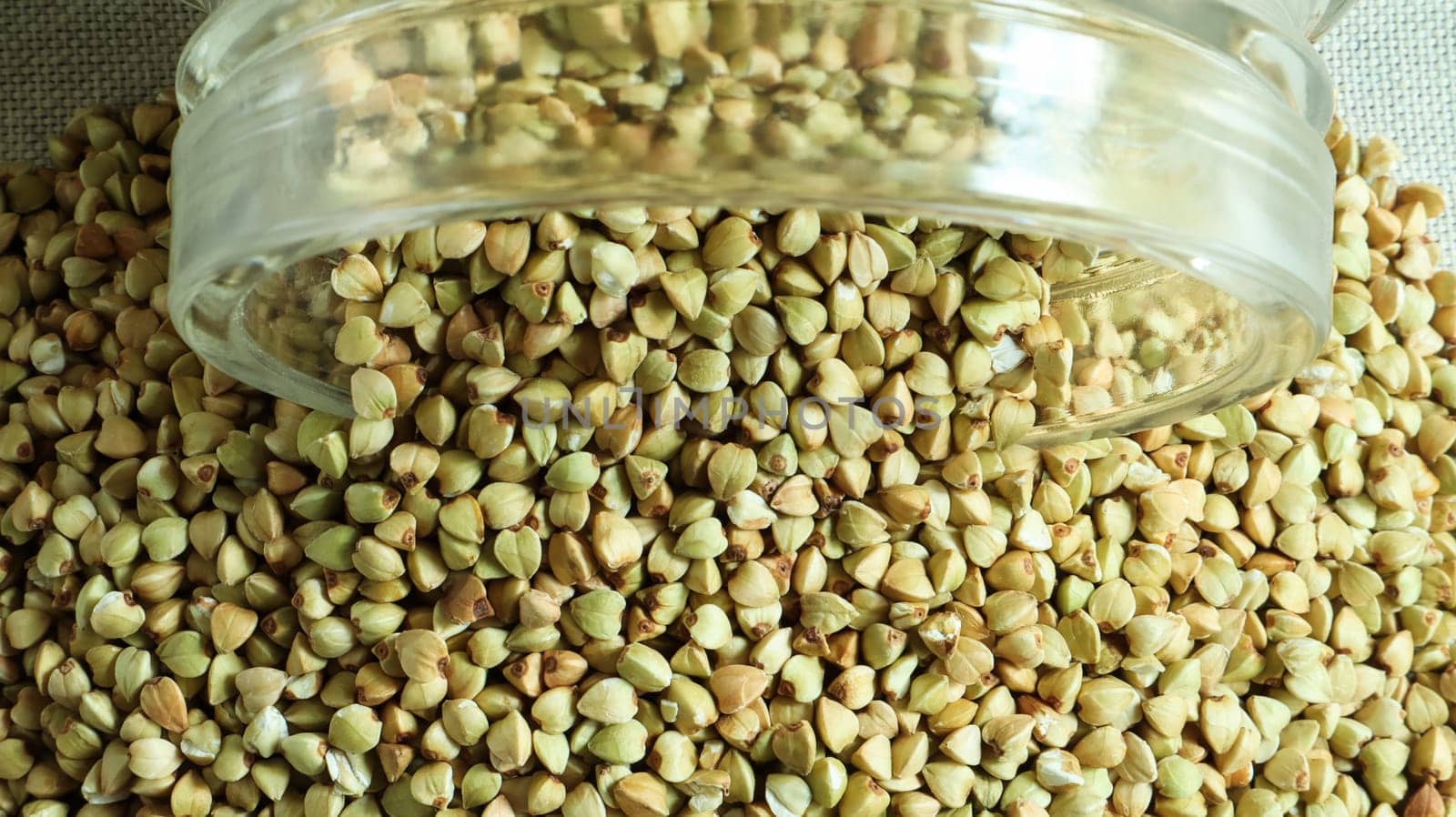 Scattered raw green buckwheat with a glass jar for cereals. Vegan organic food concept. The concept of diet, weight loss, healthy and proper nutrition. Triangle shaped scatter kernels.