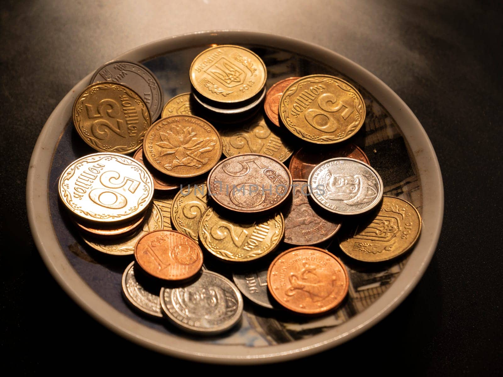 Small money coins in a plate on a black background. Eurocent. Ukrainian hryvnia. Chinese yuan. Currency unit. Small coin. Wealth. Treasure. Background image. Place for text. Poster.