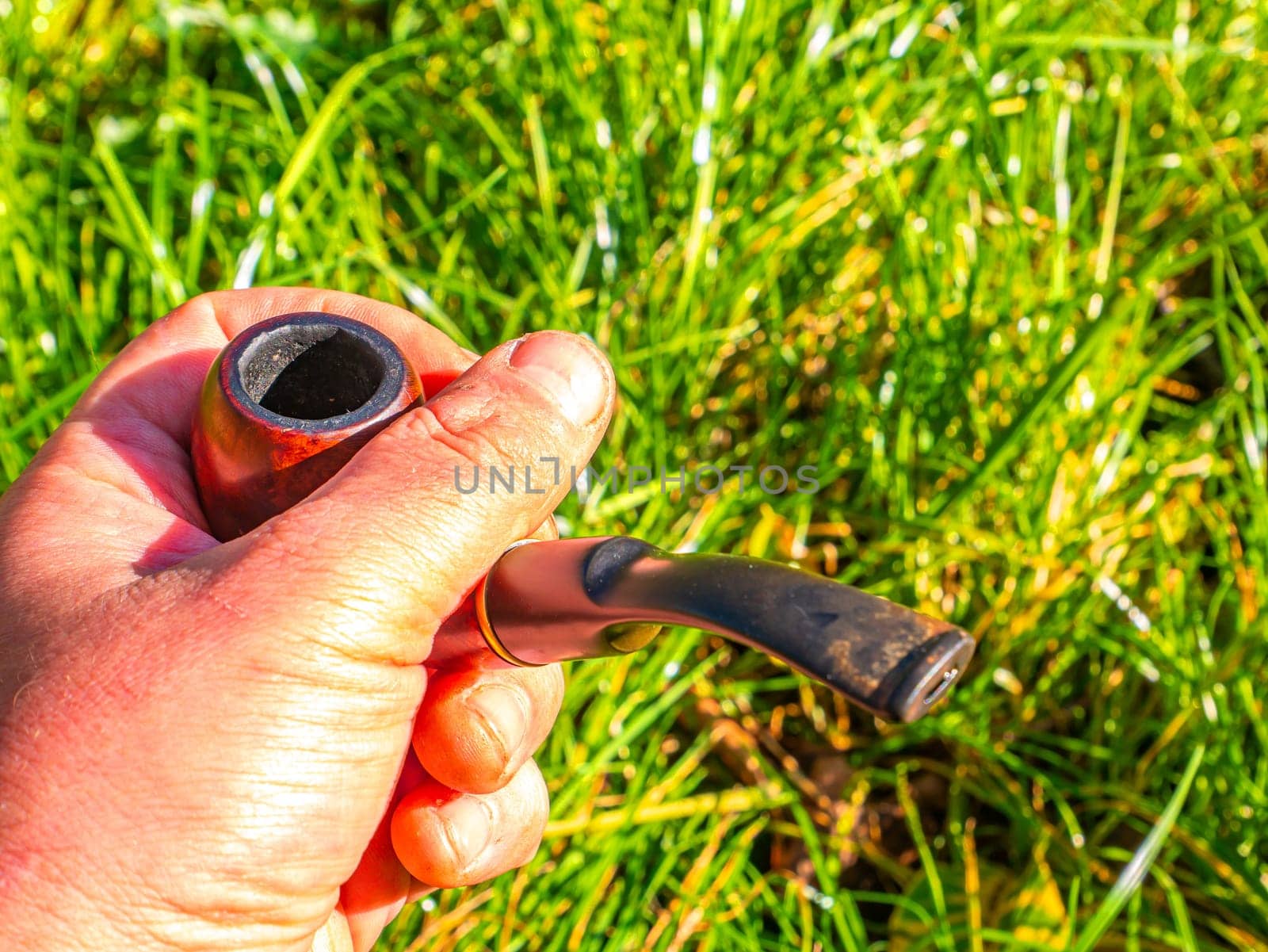 Wooden smoking pipe for tobacco in hand. Pipe for smoking tobacco. Smoker's tool. Human hand. Bad habits. Nicotine addiction. Nicotine in cigarettes. Green grass. Background image. Poster.