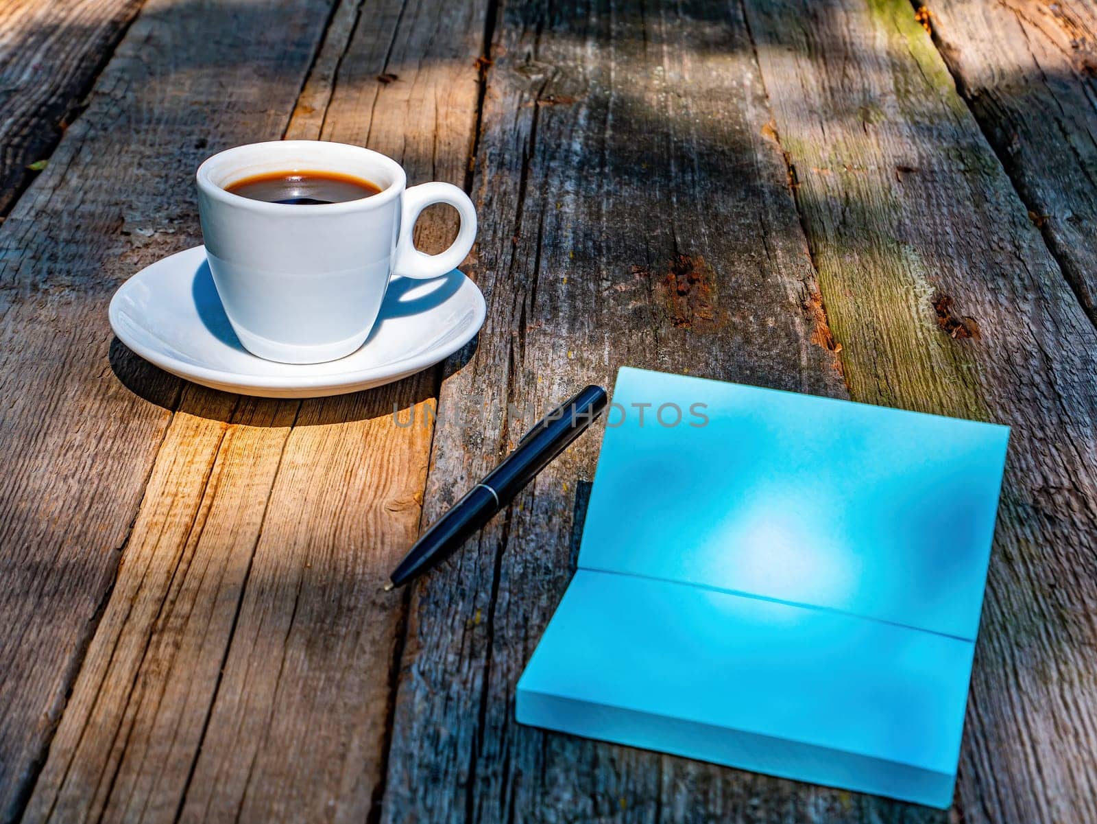 A cup of coffee and a notebook with a writing pen on a wooden tabletop. Coffee drink. Notebook. Cup and saucer. Wooden table. Food and drinks. Writing pen. Street Cafe. A business meeting.
