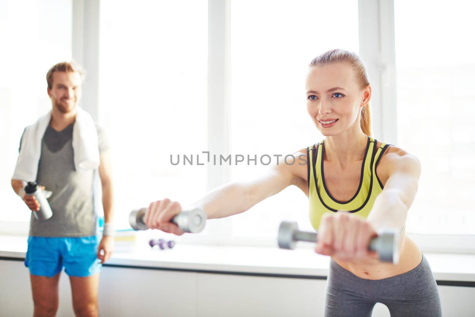 Young female exercising with dumbbells with guy on background