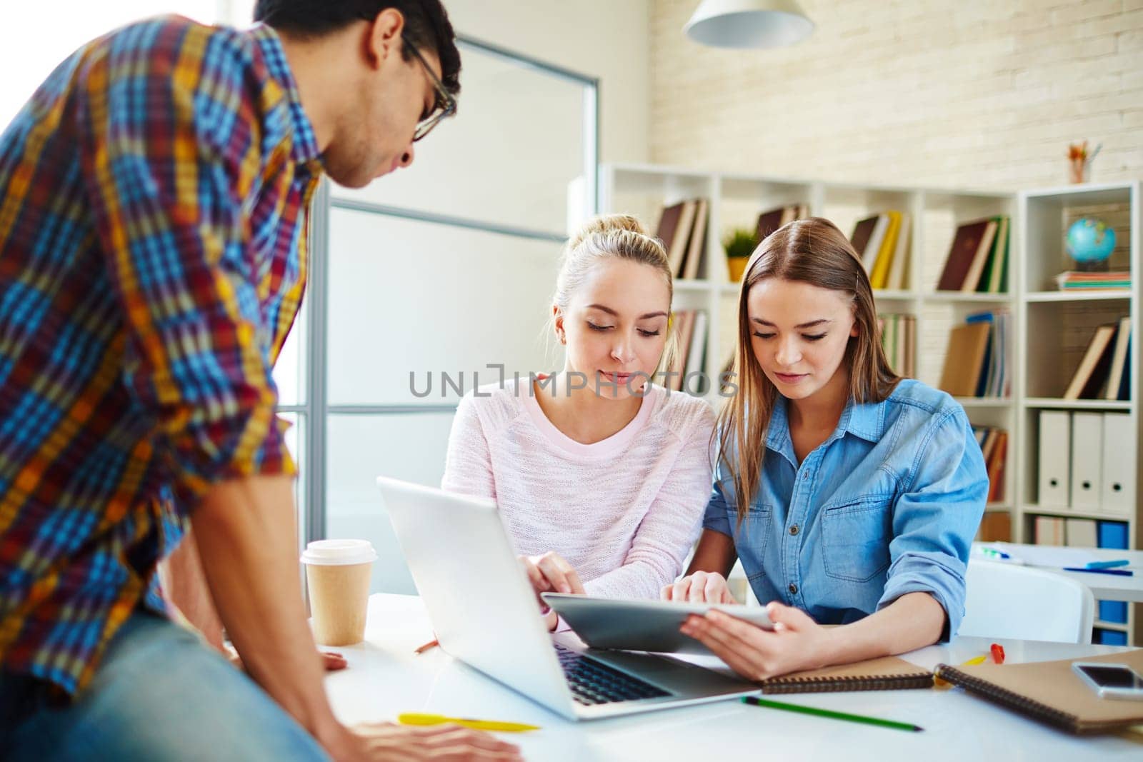 Two cute teenage girls looking through data in touchpad with their groupmate near by