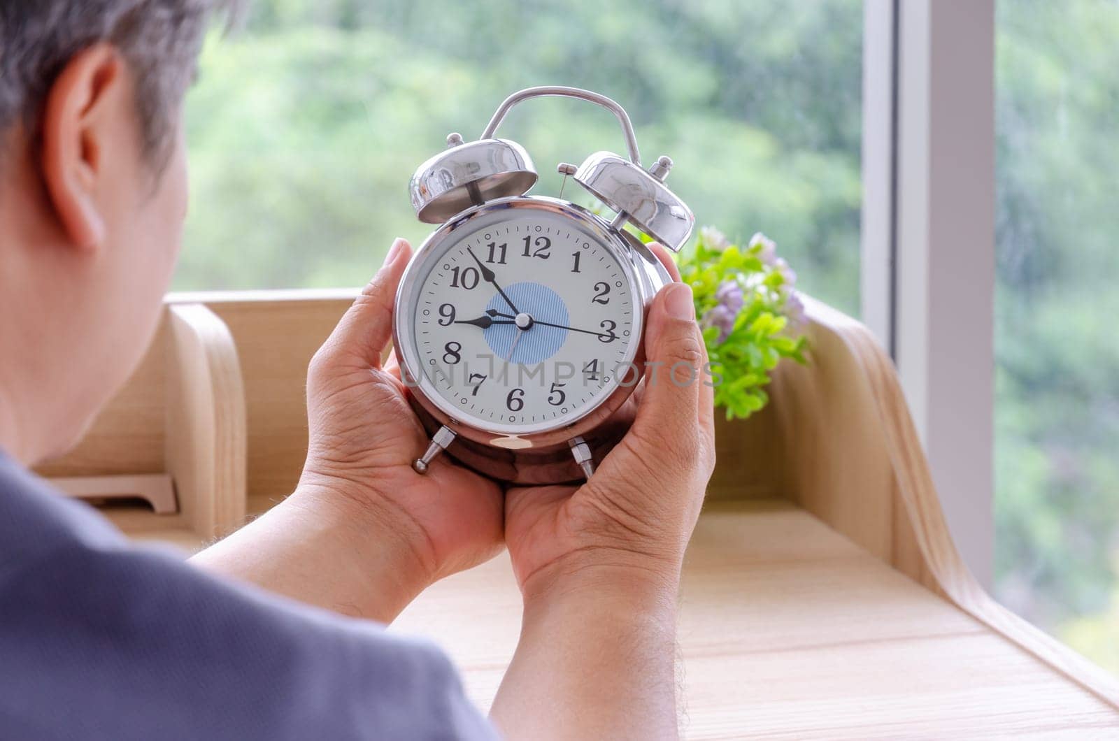 Senior man sitting at desk and holding white alarm clock