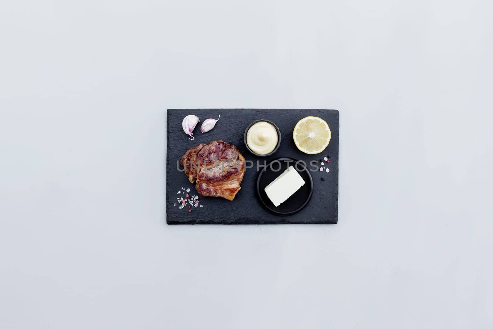 Gray concrete background flat lay with cut of medium-rare red ribeye meat steak on black cutting board, lemon, pepper seasoning, salt, mayonnaise sauce, garlic, butter. Rib-eye fillet with copy space