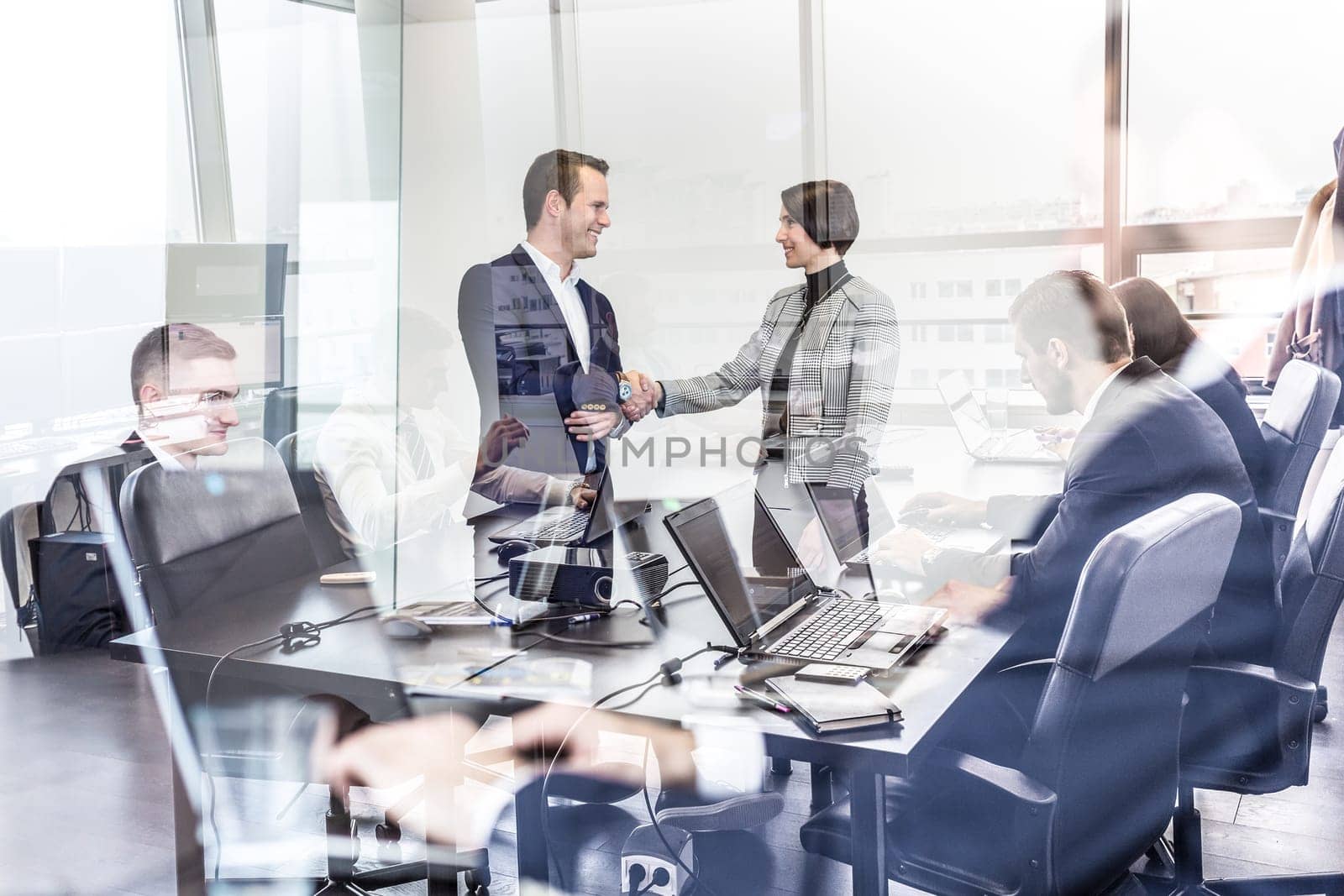 Sealing a deal. Business people shaking hands, finishing up meeting in corporate office. Businessmen working on laptop seen in glass reflection. Business and entrepreneurship concept.