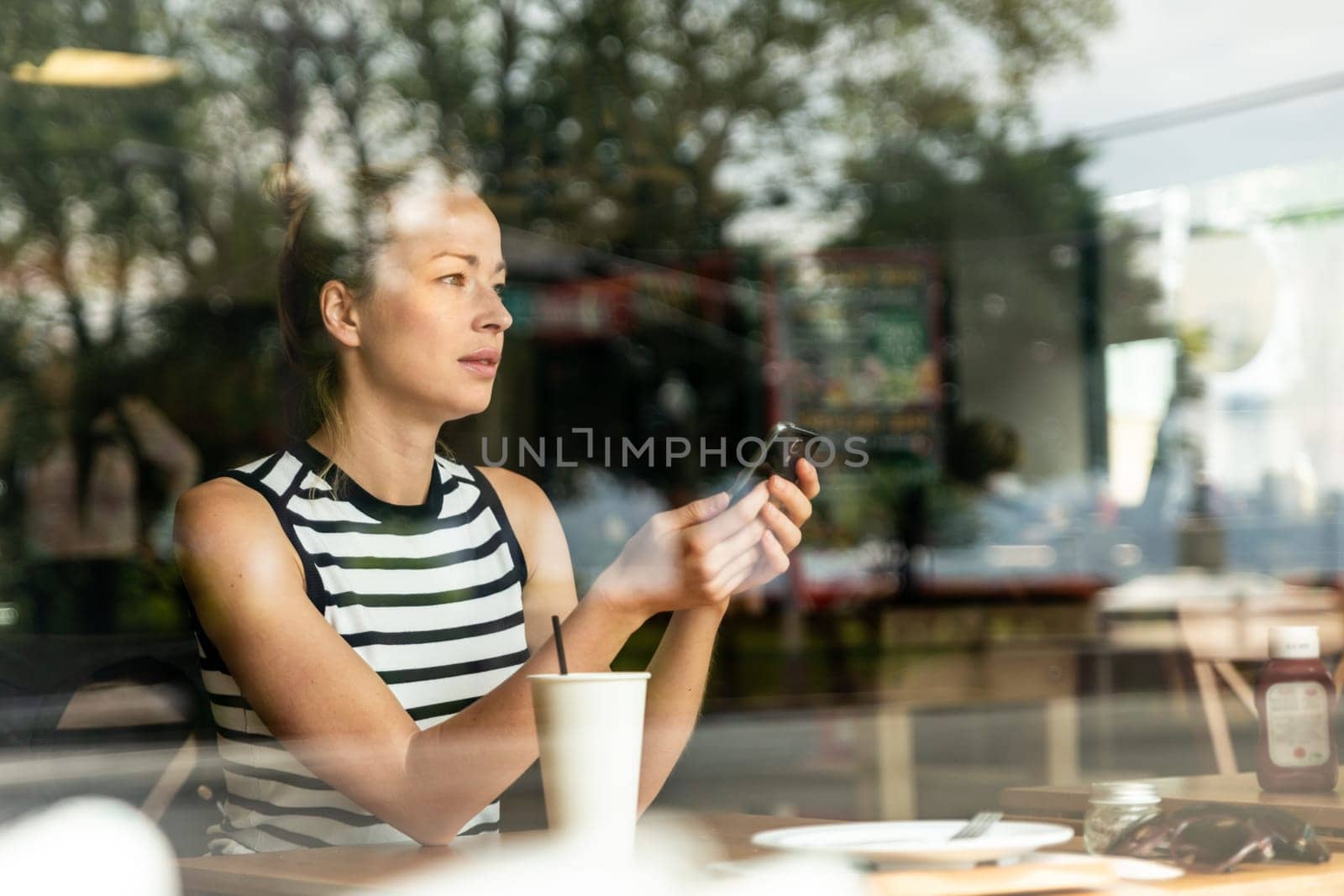 Thoughtful casual caucasian woman holding mobile phone while looking through the coffee shop window during coffee break. Street reflections in the window glass.
