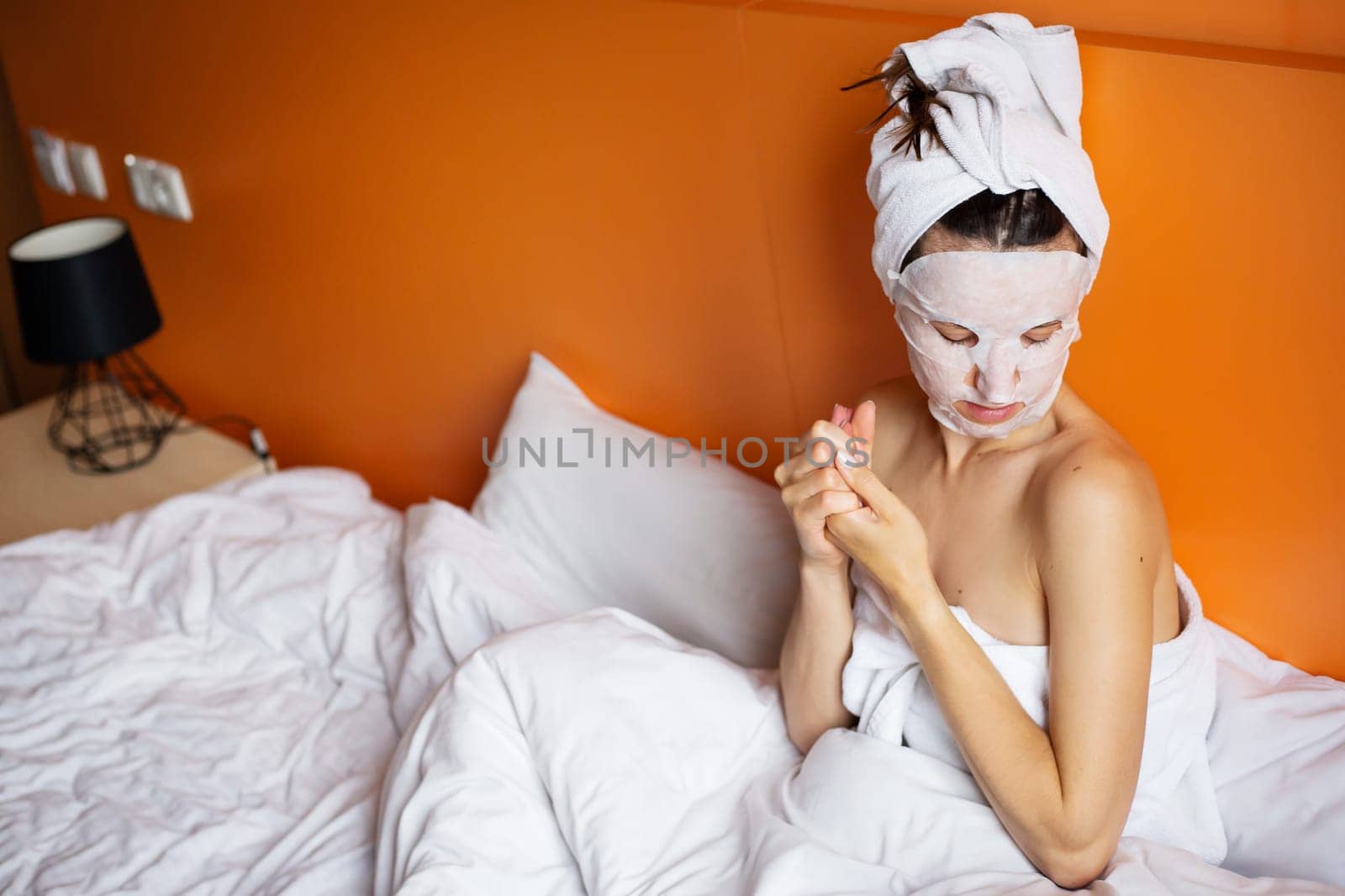 Young girl with clean skin with a mask on her face, sitting in bed, resting after a shower, relaxing at home. Close-up