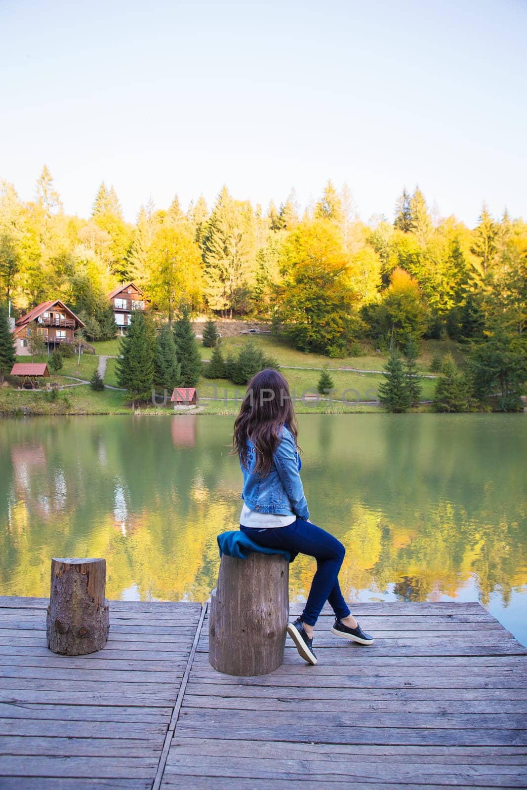 the girl is sitting and looking at the beautiful houses by the lake in the autumn forest.
