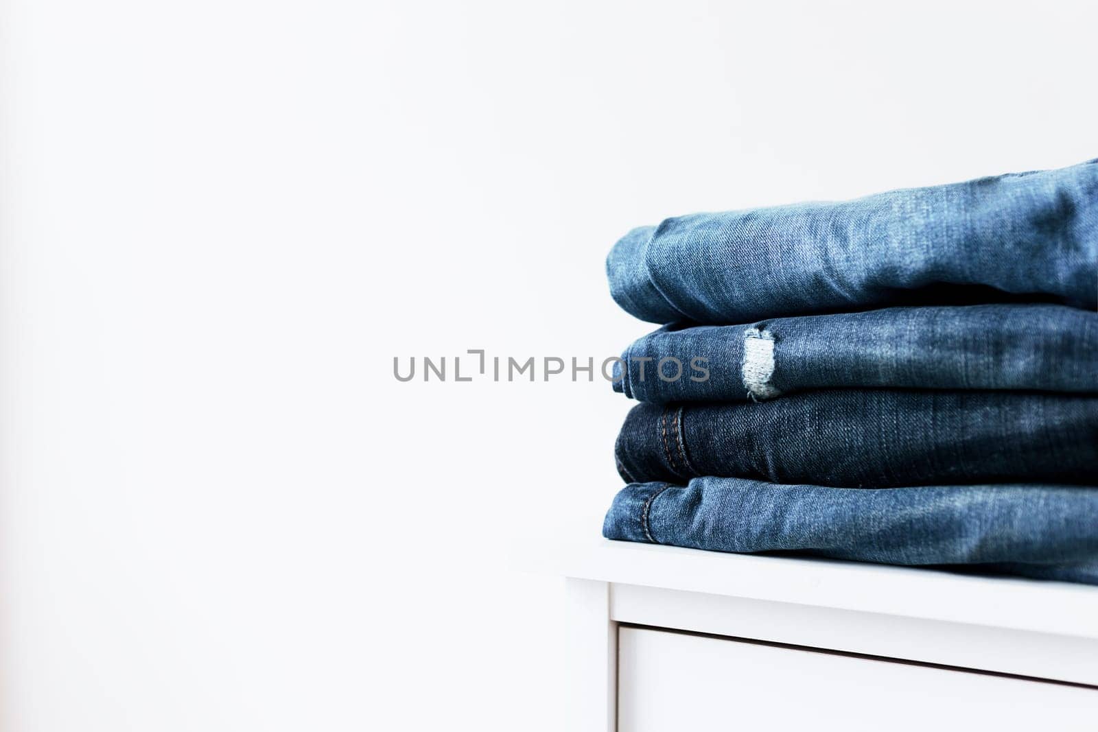 Stack of different blue denim jeans trousers on the shelf or table. Photo of stacked various shade jeans close up on white background with copy space for text design. Canvas denim fashion texture.