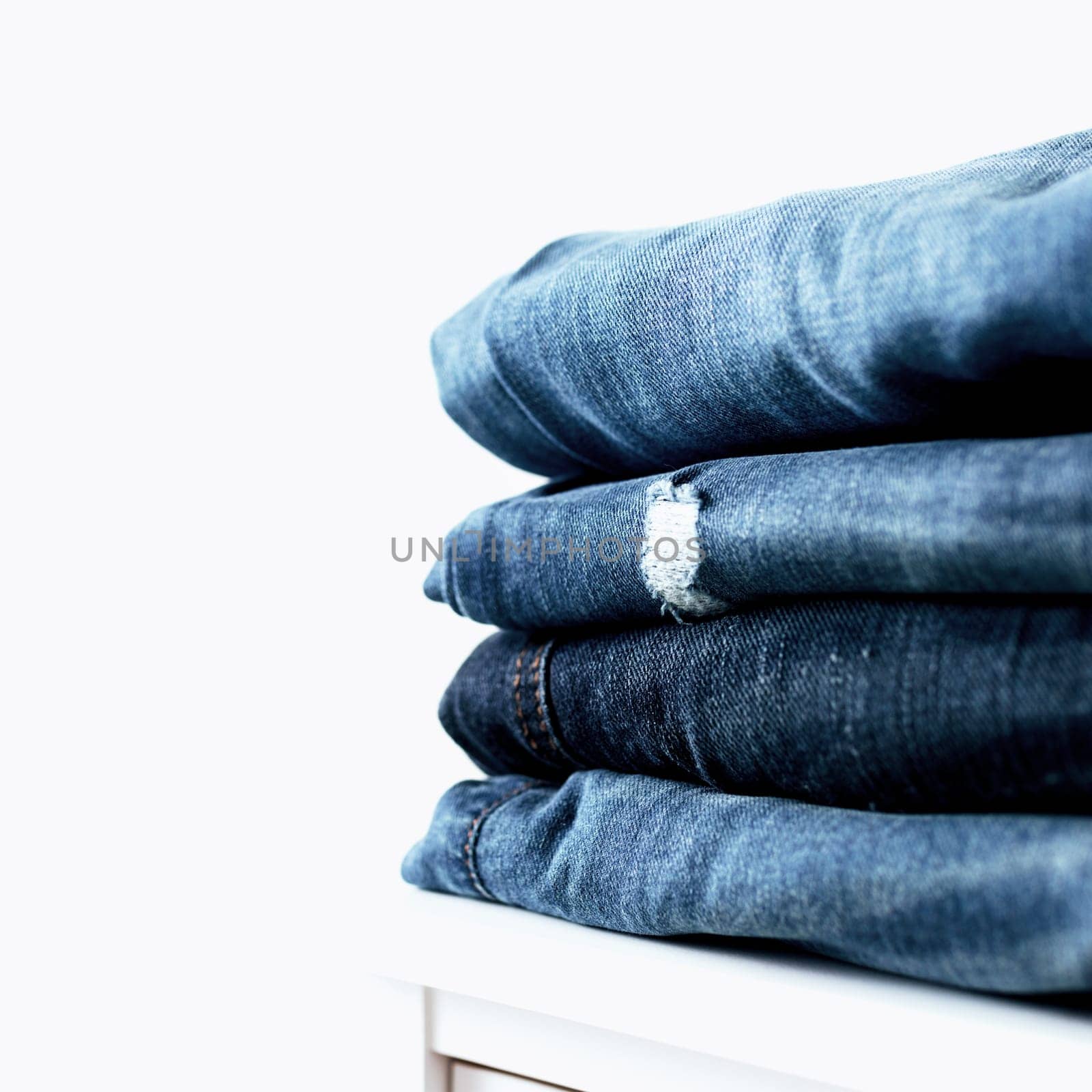 Stack of different blue denim jeans trousers on the shelf or table. Photo of stacked various shade jeans close up on white background with copy space for text design. Canvas denim fashion texture.