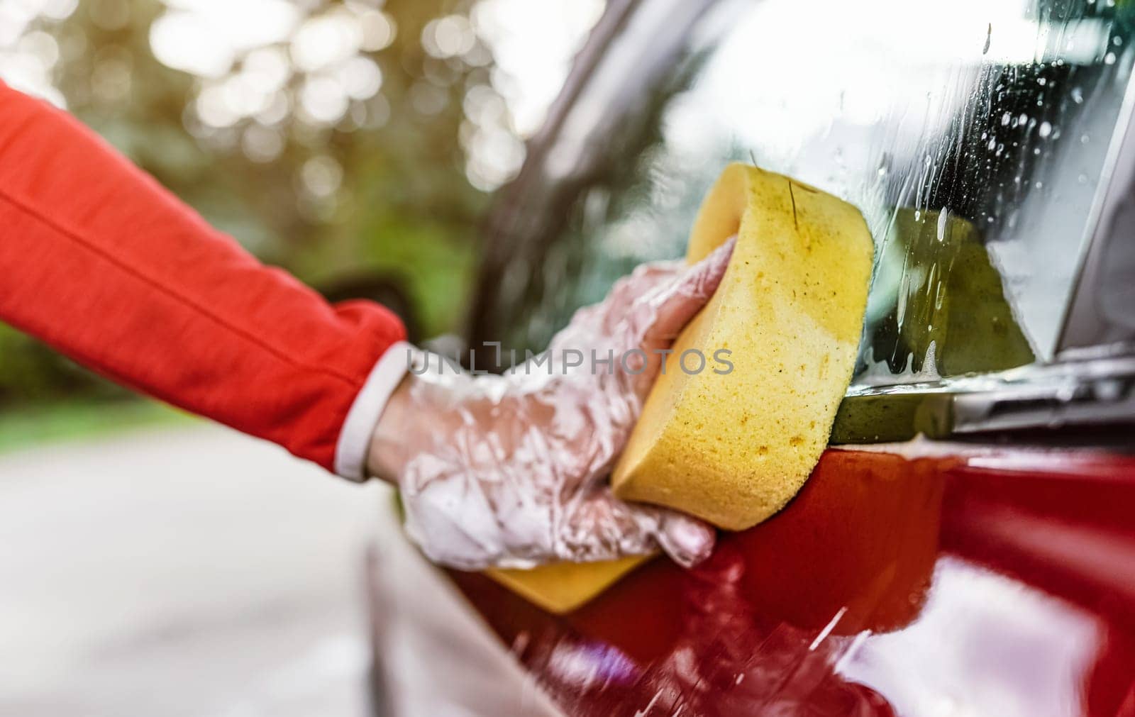 Young woman cleaning side of her car, closeup detail on hand in glove holding yellow sponge.