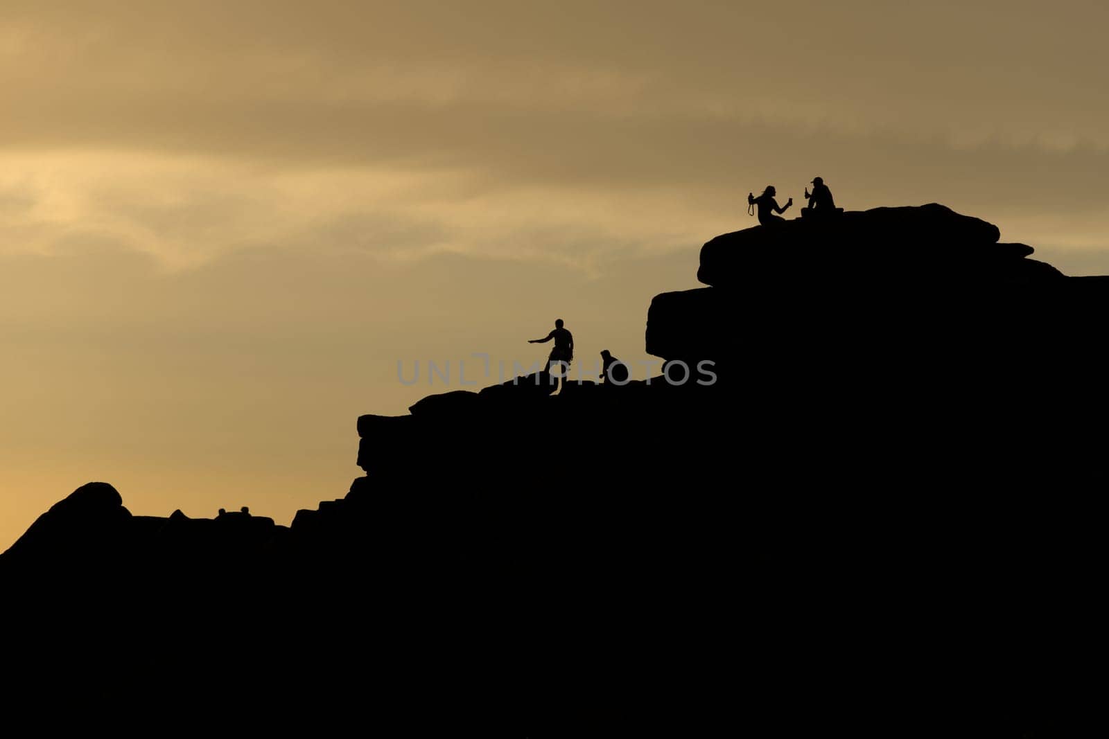 People on stanage edge, peak district, at sunset. A couple take a selfie holding drinks, silhouetted against the golden sky. Climbers stand there too