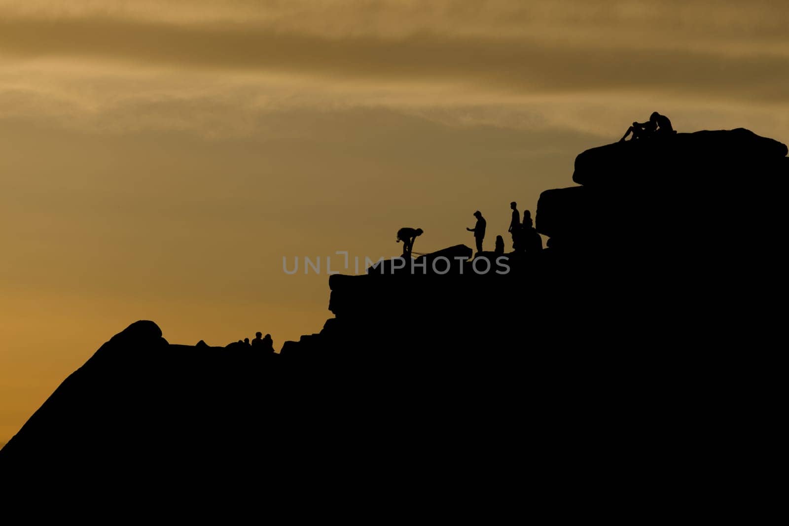 Crowds of people, including a romantic couple, surround a climber at the top of stanage edge in the peak district. Tourists enjoy the outdoors