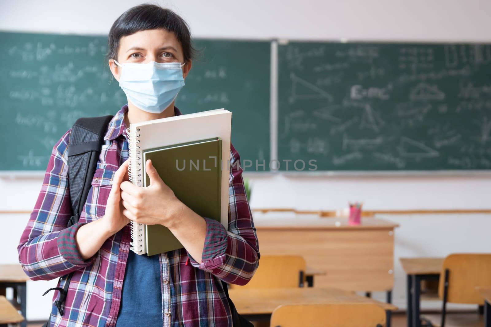 Smiling student girl wearing school backpack, mask and holding exercise book. Social distanting and classroom safety during coronavirus epidemic