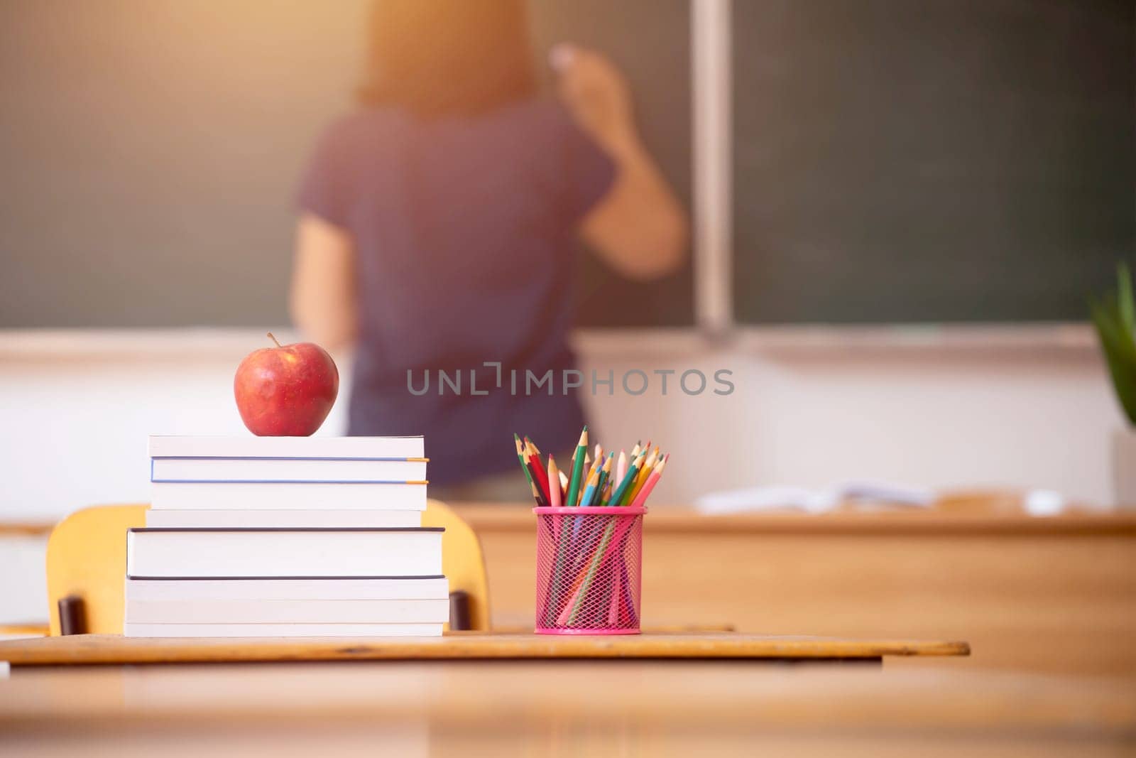 Back to School Concept. Apple, accessories and books in the classroom, chalkboard and teacher teaching in the background.