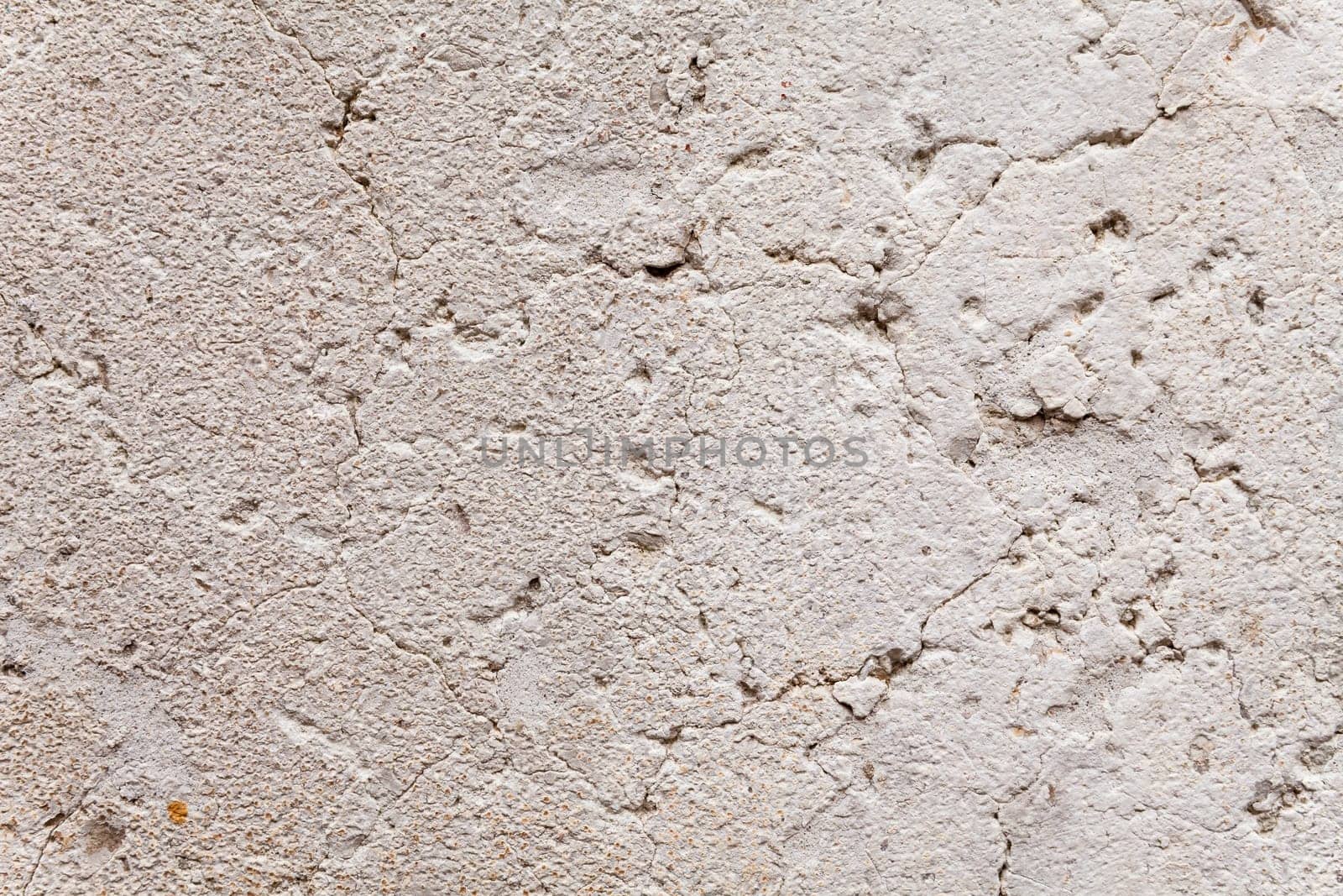 Close up on opened-up cracks in the weathered plaster of an ancient exterior wall. Rough, porous stone surface with aging patina. Beige and gray color with rust brown freckles. Texture for background.