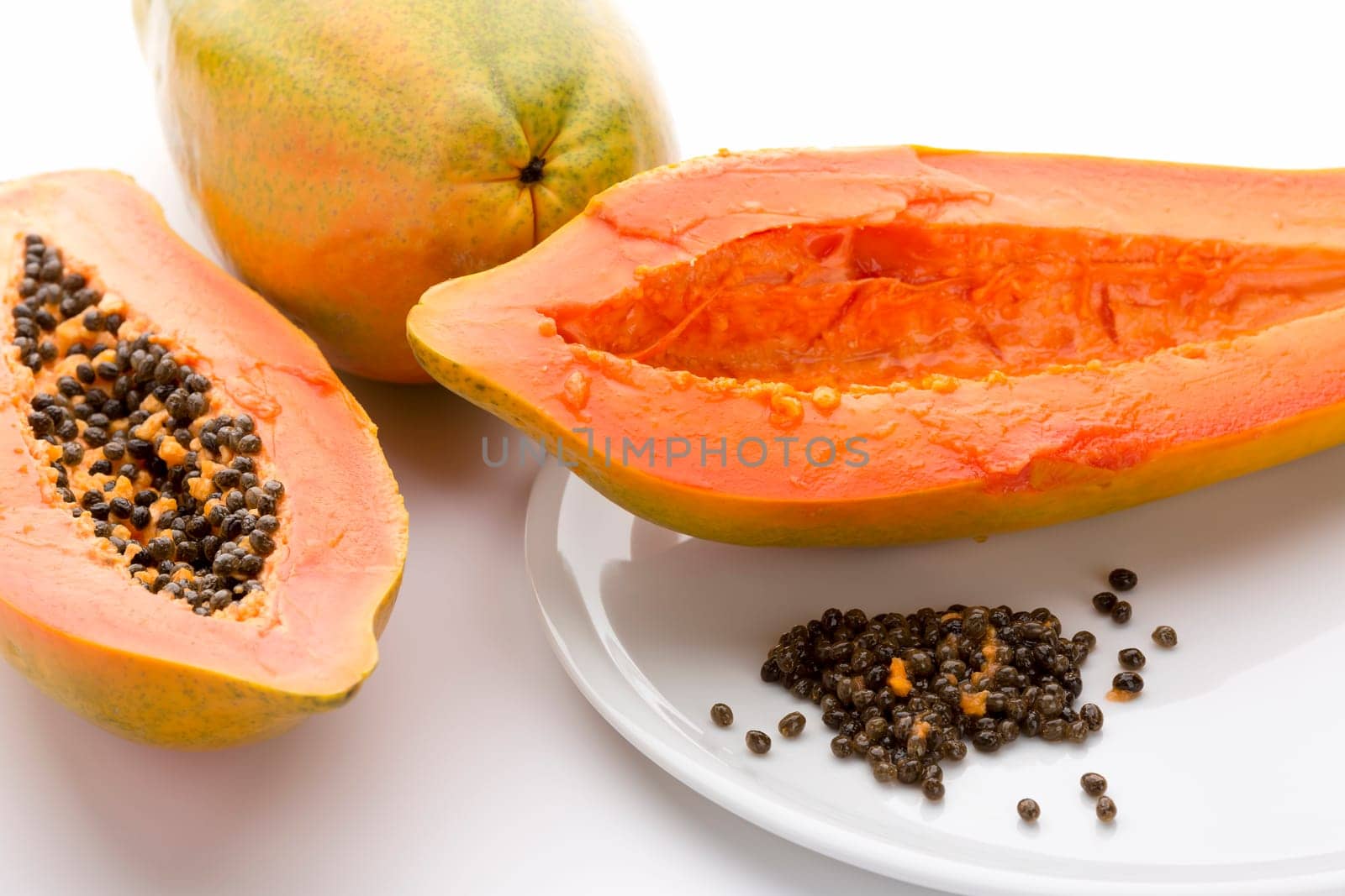 Hollowed out half of a raw papaya in longitudinal cross section and its countless peppery seeds on a white plate. Succulent breakfast fruit of the tropical Carica papaya. Close up. White background.
