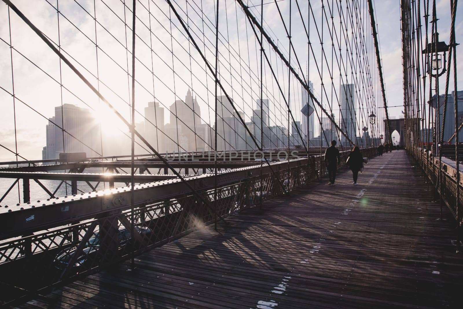 Couple walking on pedestrian path across Brooklyn bridge. New York City Manhattan downtown skyline in sunset with skyscrapers illuminated over East River panorama as seen from Brooklyn bridge.