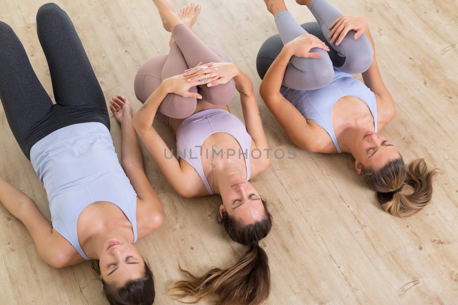 Group of three young sporty attractive women in yoga studio, lying on the floor, stretching and relaxing after the workout. Healthy active lifestyle, working out indoors in gym.