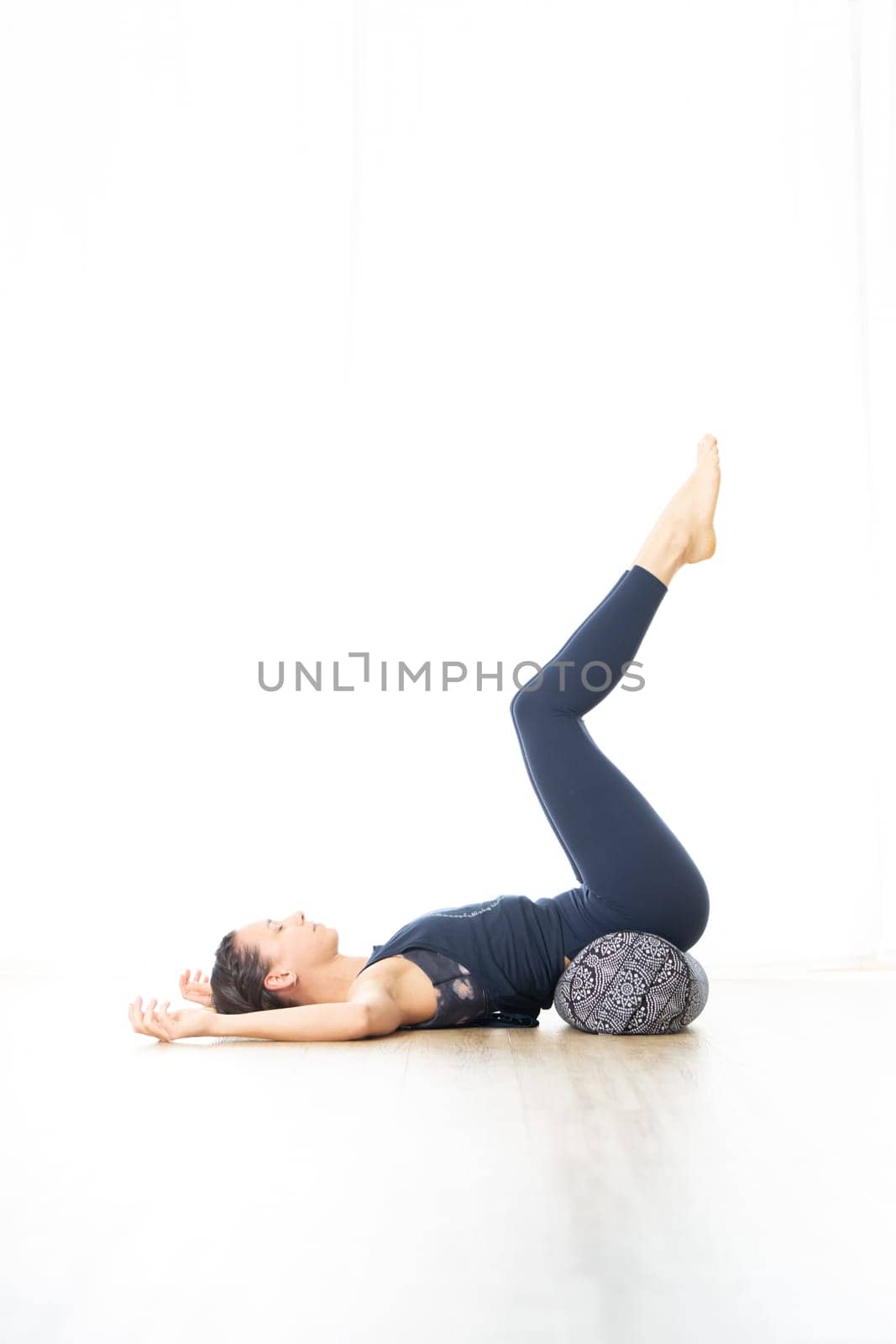 Restorative yoga with a bolster. Young sporty attractive woman in bright white yoga studio, lying on bolster cushion, stretching and relaxing during restorative yoga. Healthy active lifestyle.