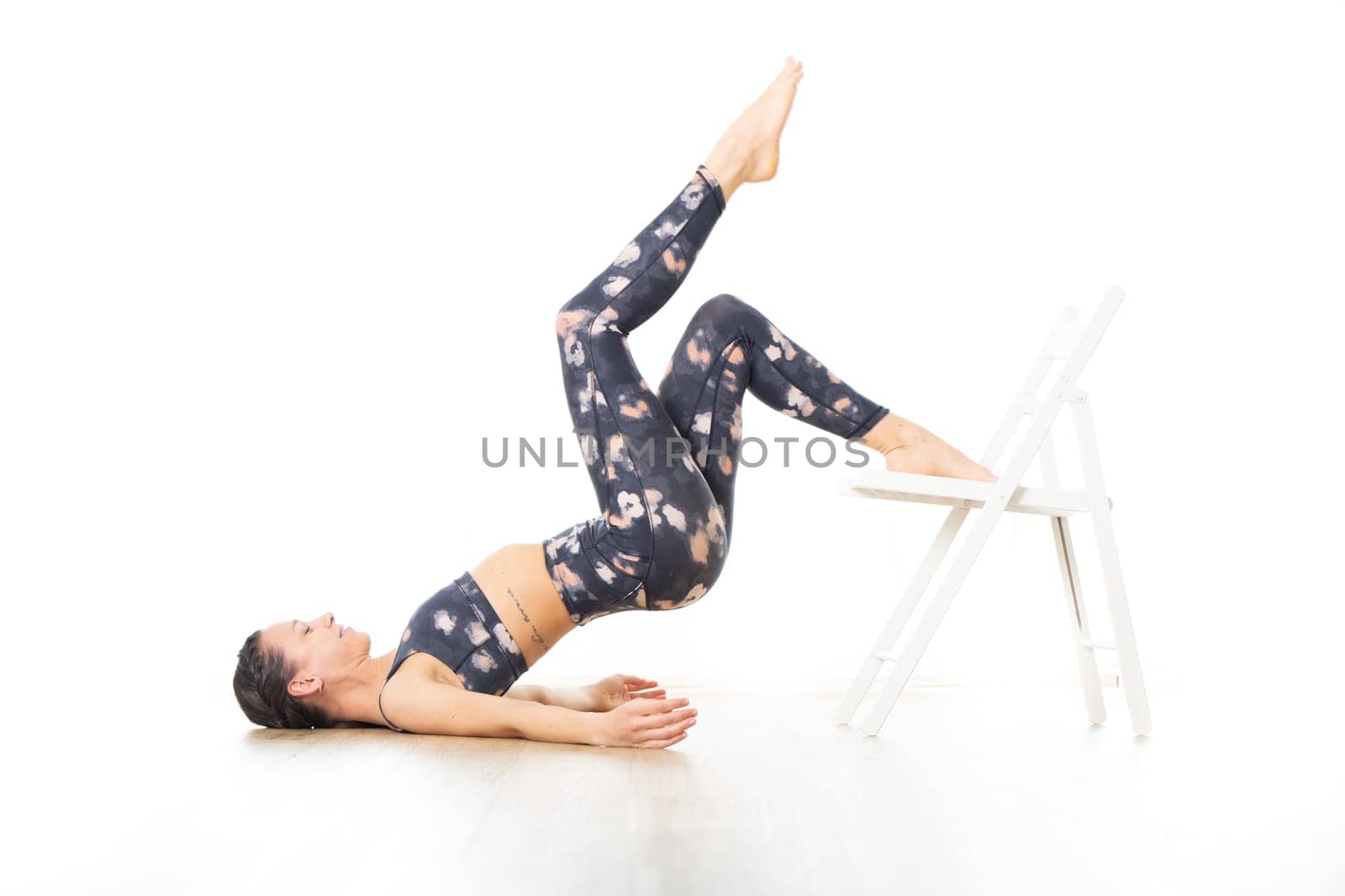 Young sporty attractive woman in bright white yoga studio, stretching and relaxing during restorative yoga using white chair as a gadget. Healthy active lifestyle.
