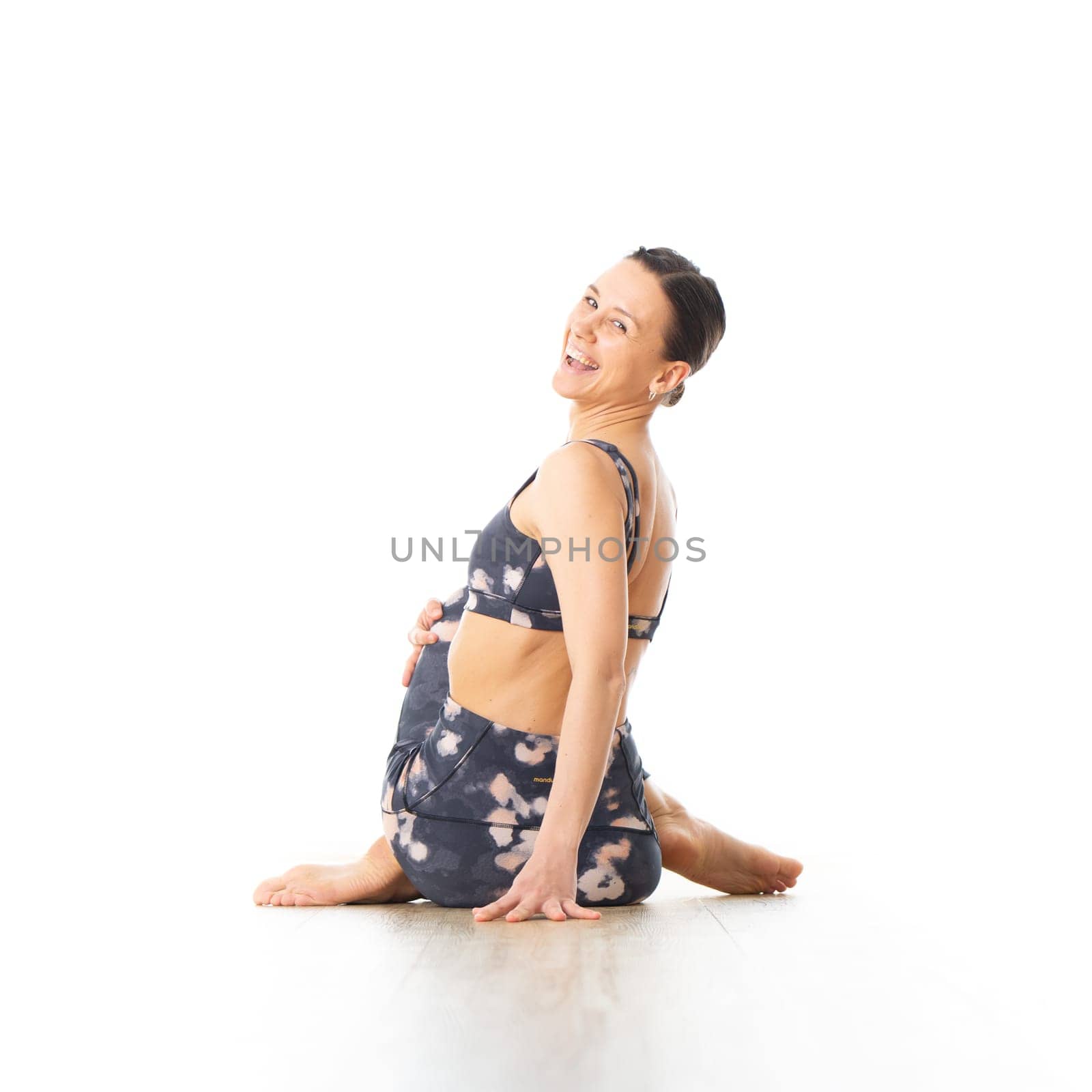 Rear view of young sporty female yoga instructor in bright white yoga studio, smiling cheerfully while looking in camera