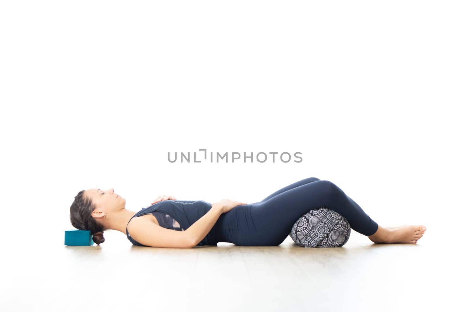 Restorative yoga with a bolster. Young sporty attractive woman in bright white yoga studio, lying on bolster cushion, stretching and relaxing during restorative yoga. Healthy active lifestyle.