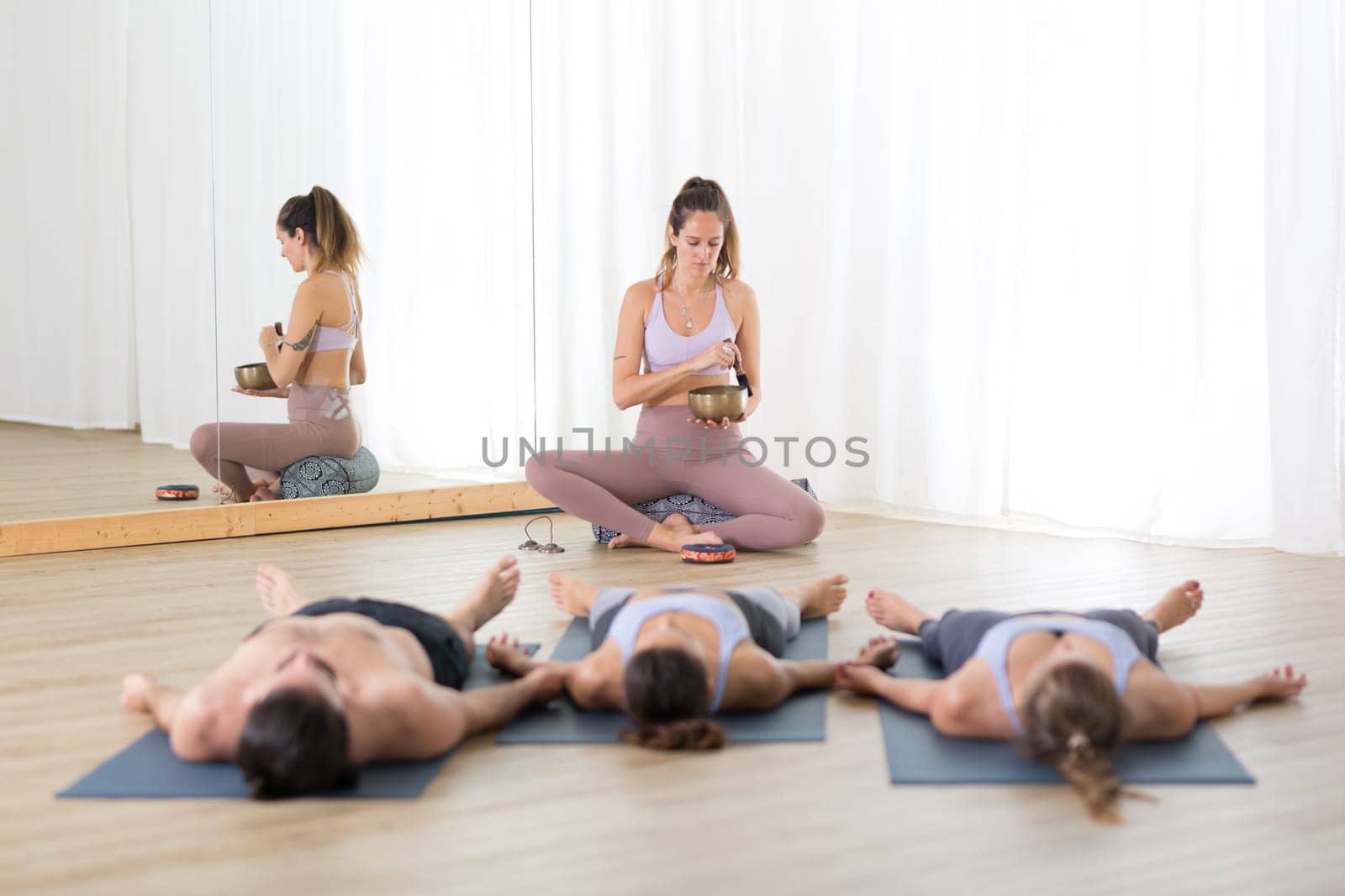 Yoga instructor and group of young sporty people in yoga studio, lying and relaxing on yoga mats during restorative yoga session. Healthy active lifestyle.