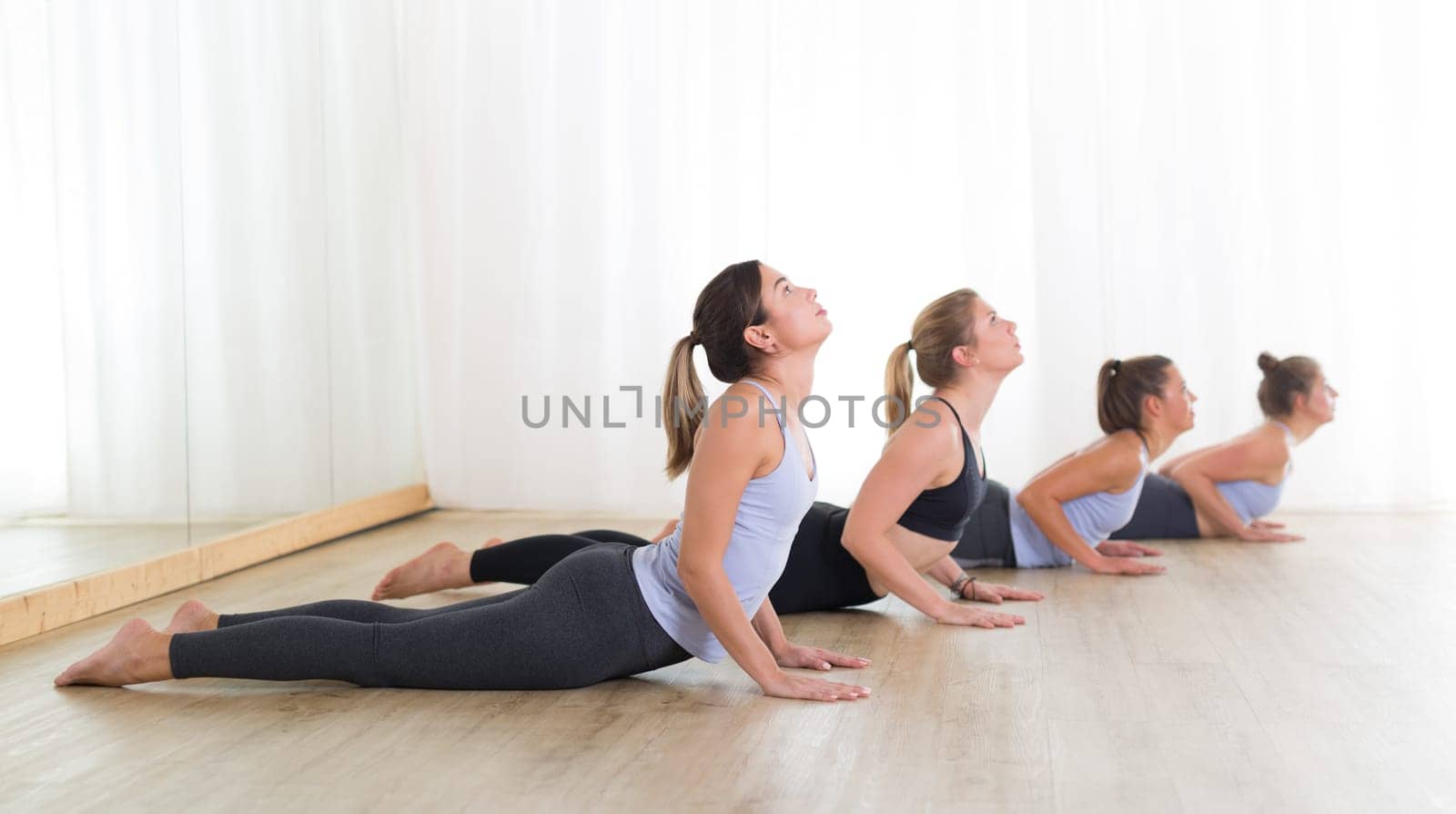 Group of young sporty sexy women in yoga studio, practicing yoga lesson with instructor, forming a line in asana pose. Healthy active lifestyle, working out indoors in gym.