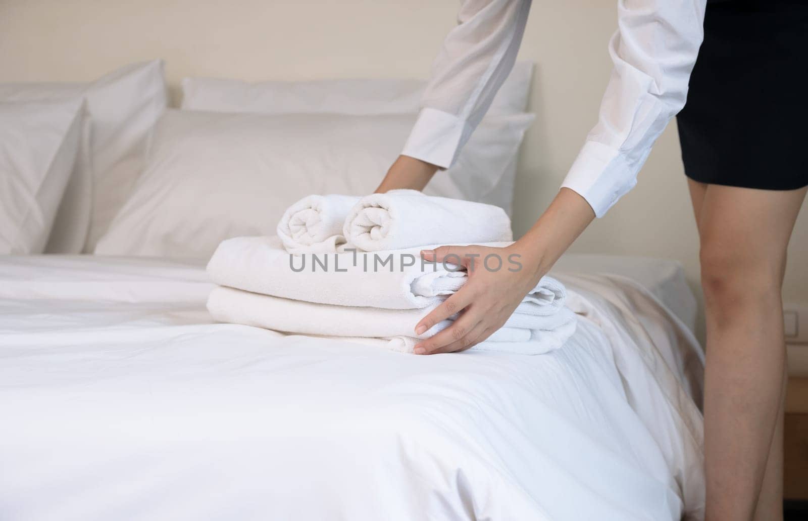 Hotel room service making room. Woman chambermaid keeping stack of fresh white bath towels on bed in guest room