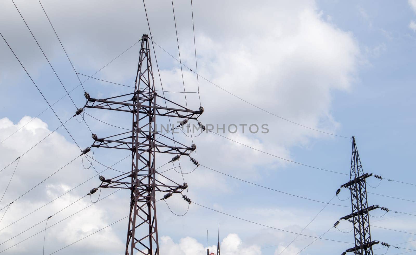 High voltage electrical tower. Power lines and power pylons on a sunny day with circular clouds in the blue sky. High voltage power transmission line supports. They have a complex steel structure