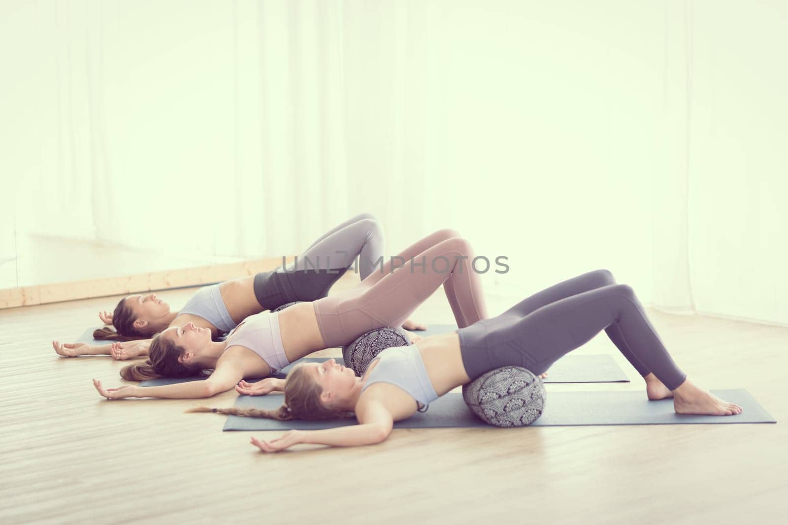 Restorative yoga with a bolster. Group of three young sporty attractive women in yoga studio, lying on bolster cushion, stretching and relaxing during restorative yoga. Healthy active lifestyle.