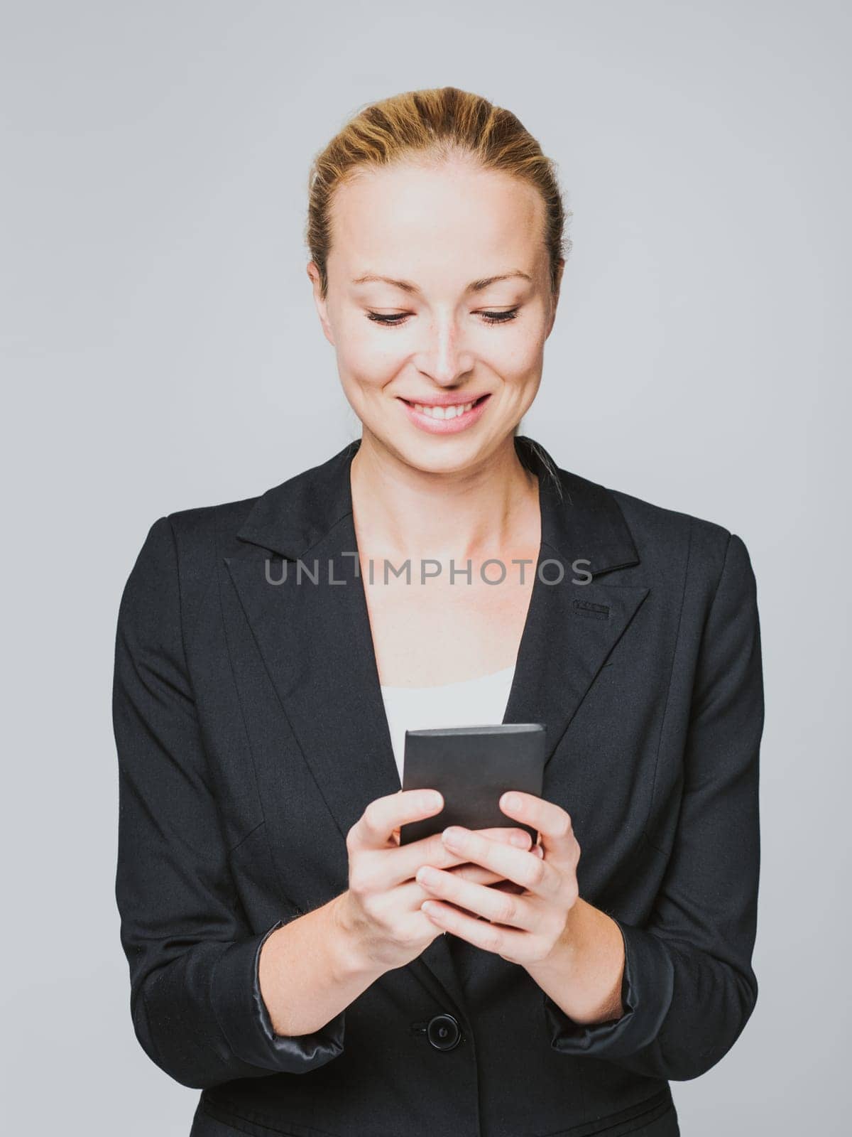 Beautiful young caucasian businesswoman in business attire using smart phone application. Studio portrait shot on gray background.