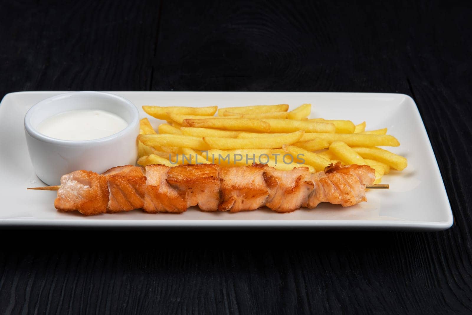 Grilled salmon fish shashlik with fried potatoes and sauce on white plate on black wooden background
