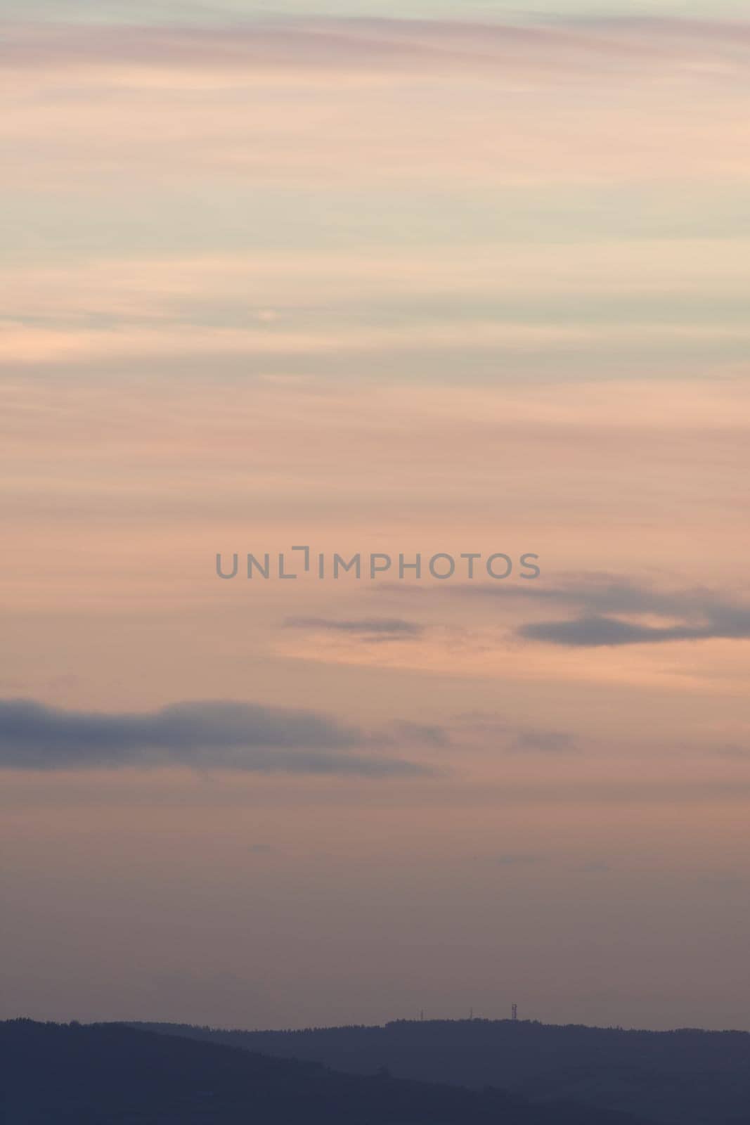 Vertical hazy sunset. Horizon under warm and faded pastel colours. Gently clouded sky above the forested horizon with protruding telecom towers.