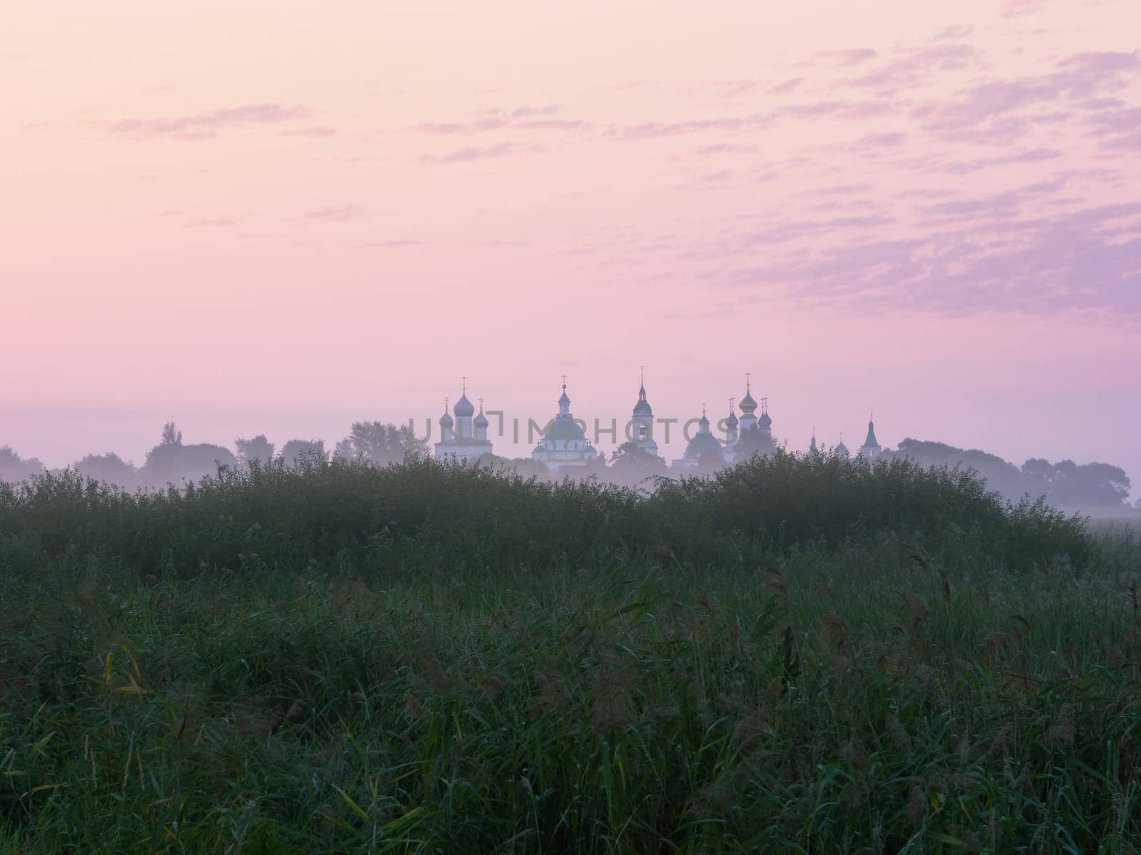Lilac-pink sky at dawn before sunrise. A distant view of the domes of an Eastern Orthodox monastery in the morning fog behind a grassy hill. Russia, the Rostov's outskirts from the side of the Monastery of St. Jacob Saviour (Spaso-Yakovlevsky).