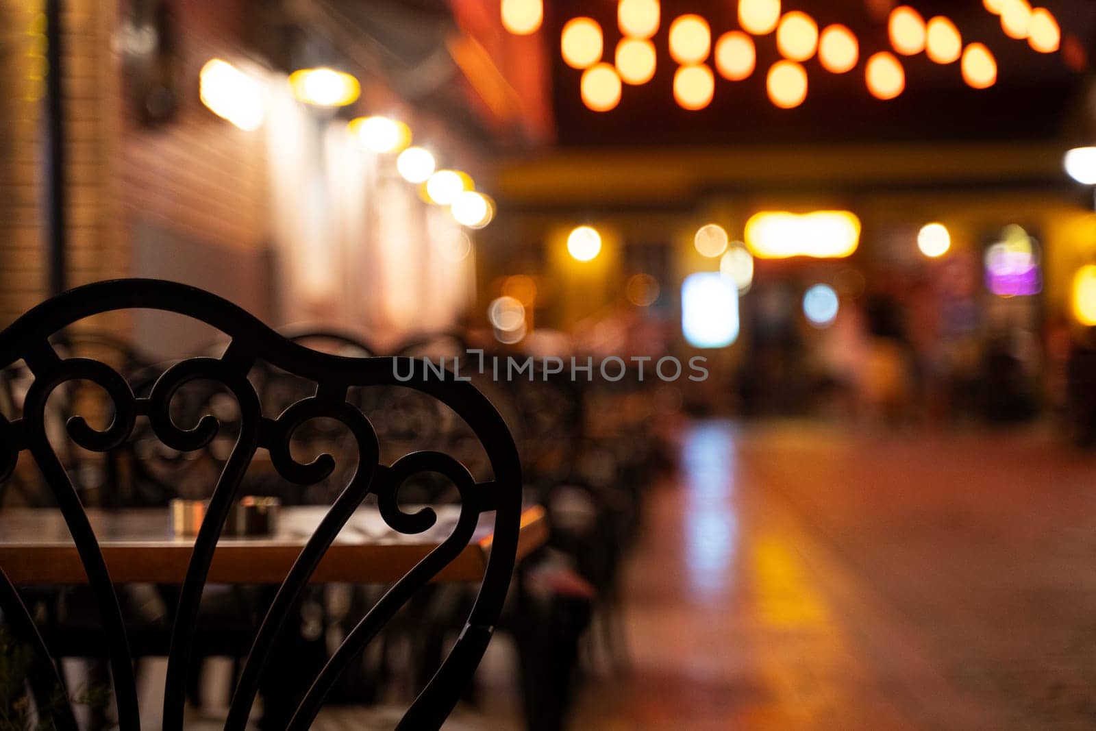 closeup rear view of black chairs with tables on restaurant terrace on a illuminated street at night with lights in the background