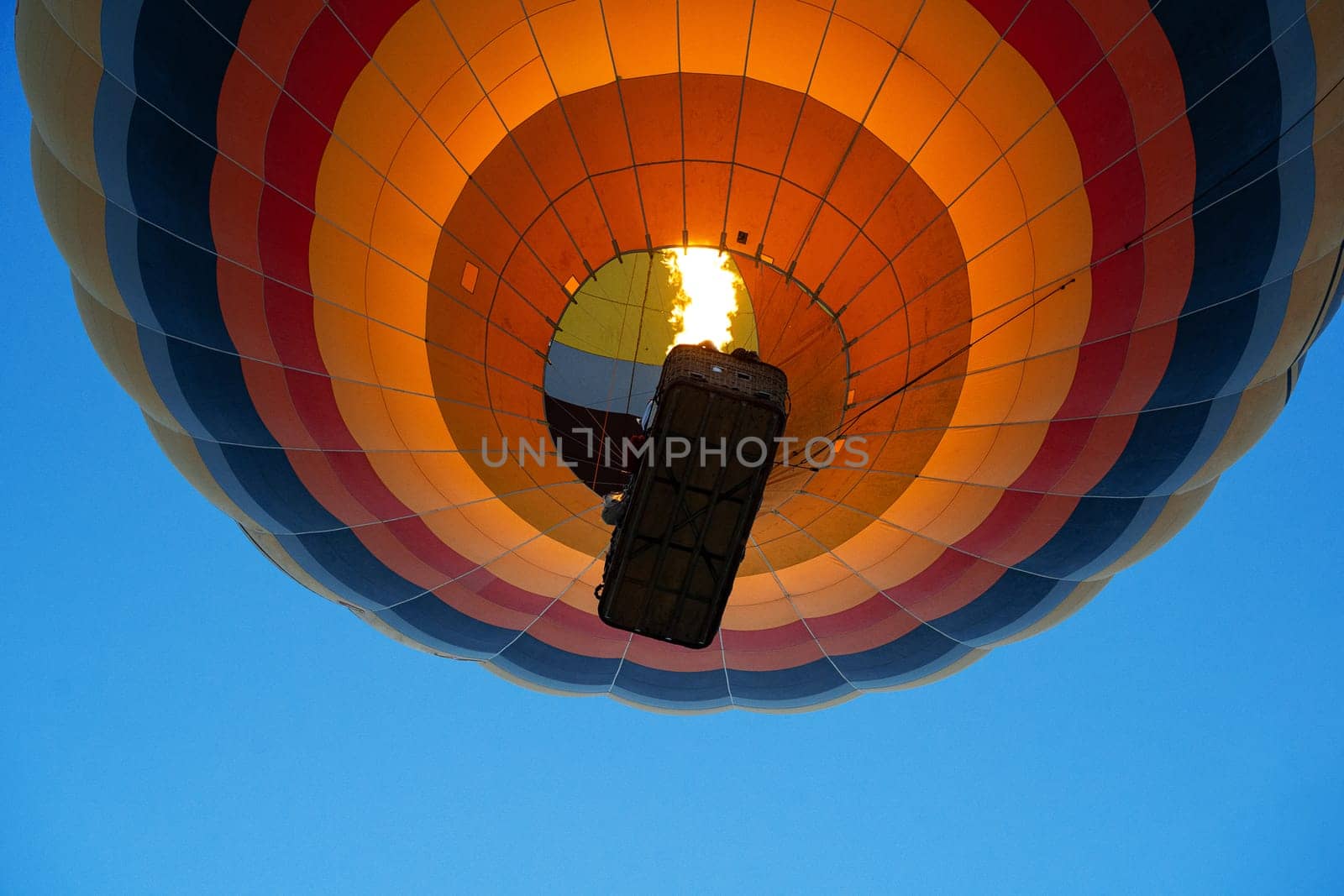 view from the ground of colorful hot air balloon with large wicker passenger basket rising up in the blue sky using burner fire flame