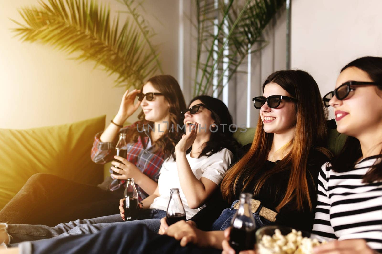 Friend group of people in 3D glasses are watching a movie, smiling, laughing. Eating and drinking while watching a movie in the cinema. Girls eat popcorn and drink soda in glass bottles. Relaxing time