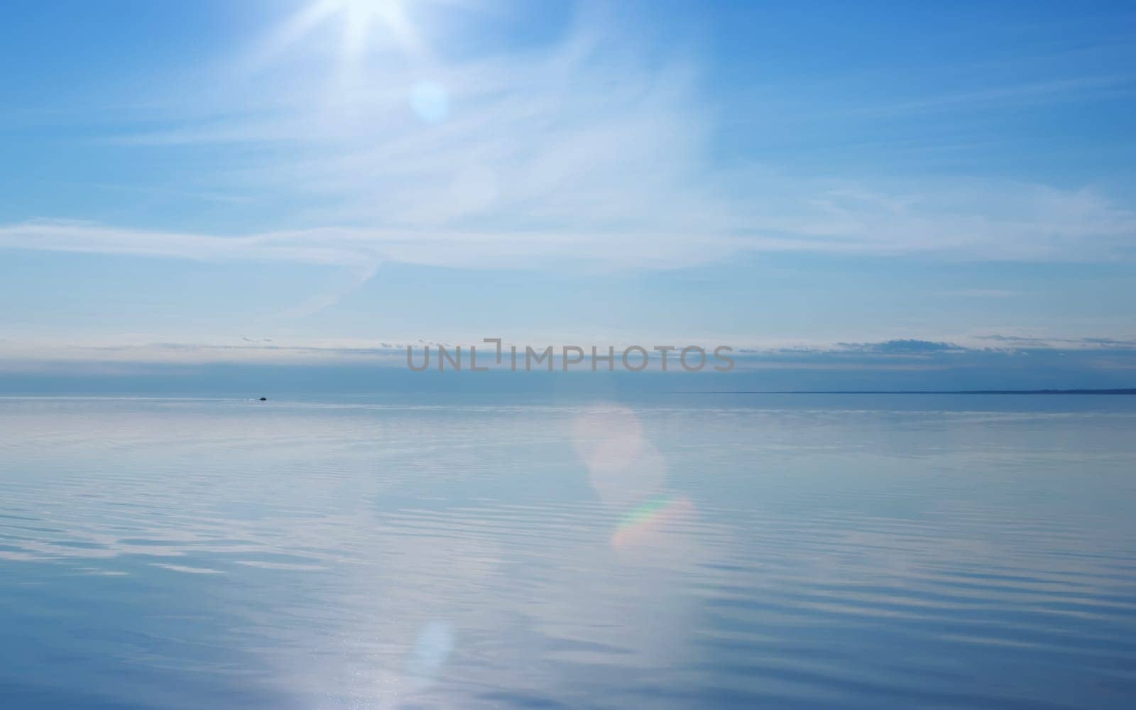 Blue sky reflected in a smooth lake surface. Water glares and shines in the rays of the evening sun with lens flare. Summer seascape background with space for copy. Russia, Vologda Oblast, Lake Beloye.