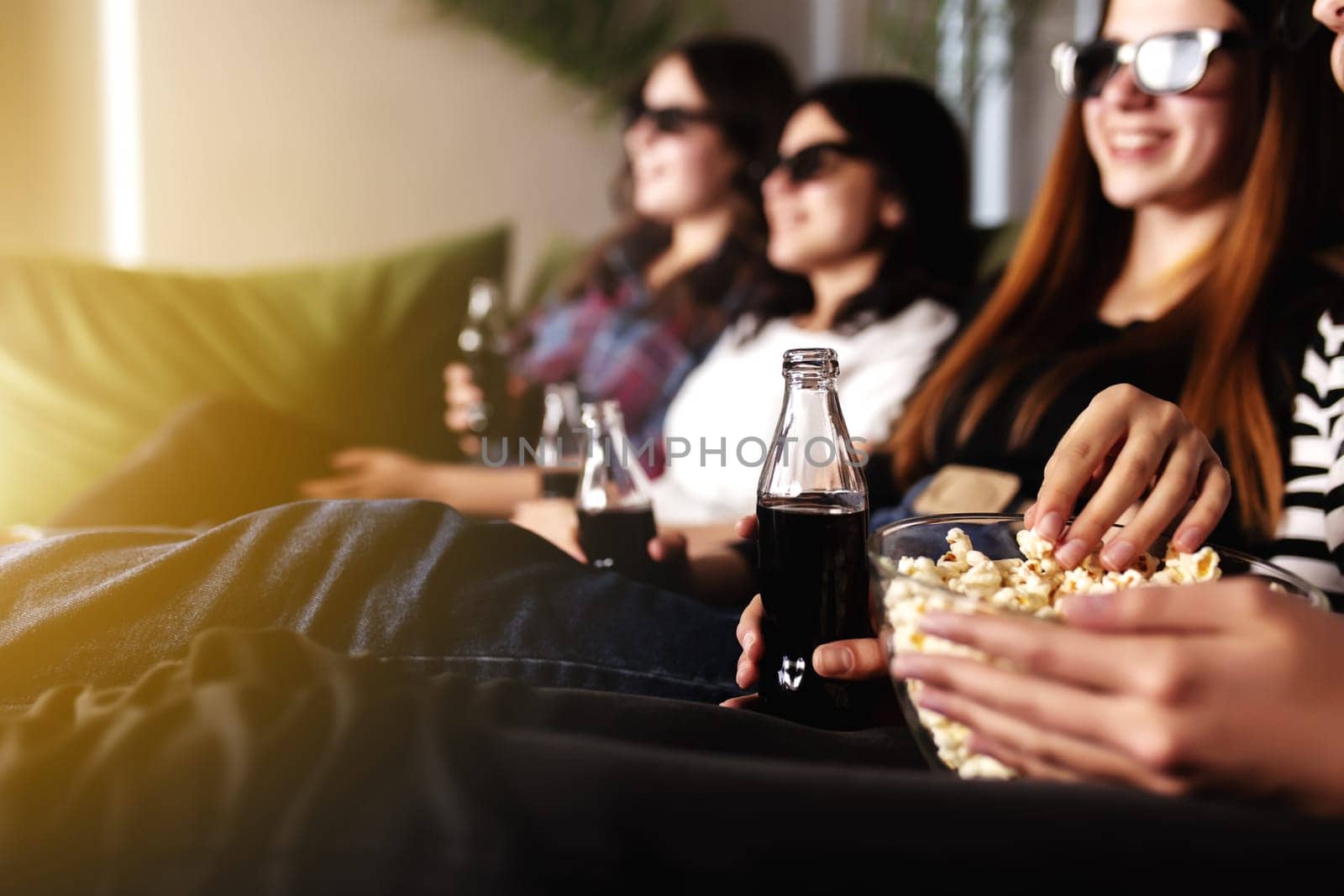 Friend group of people in 3D glasses are watching a movie, smiling, laughing. Eating and drinking while watching a movie in the cinema. Girls eat popcorn and drink soda in glass bottles. Relaxing time