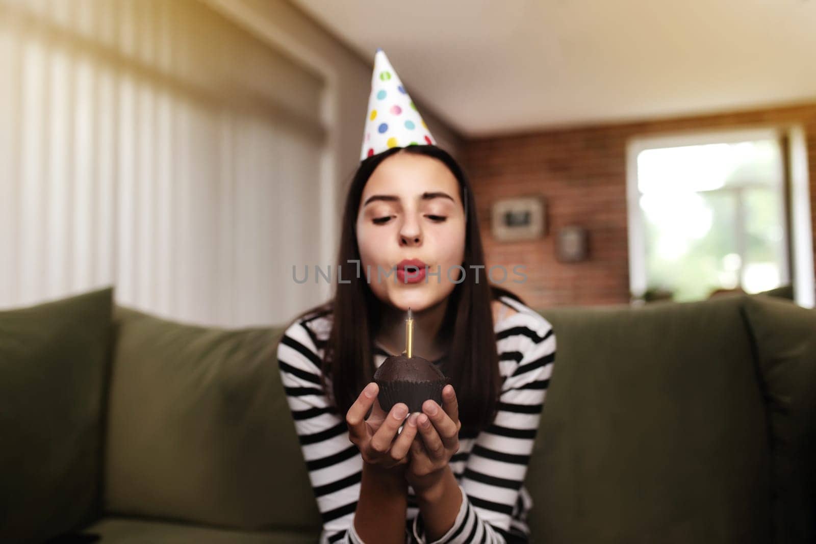 Birthday girl brunette in cardboard hat sits on the couch or sofa and blows out burning candle on cupcake. Woman holding birthday cake in her hands. Congratulation concept with blurred background